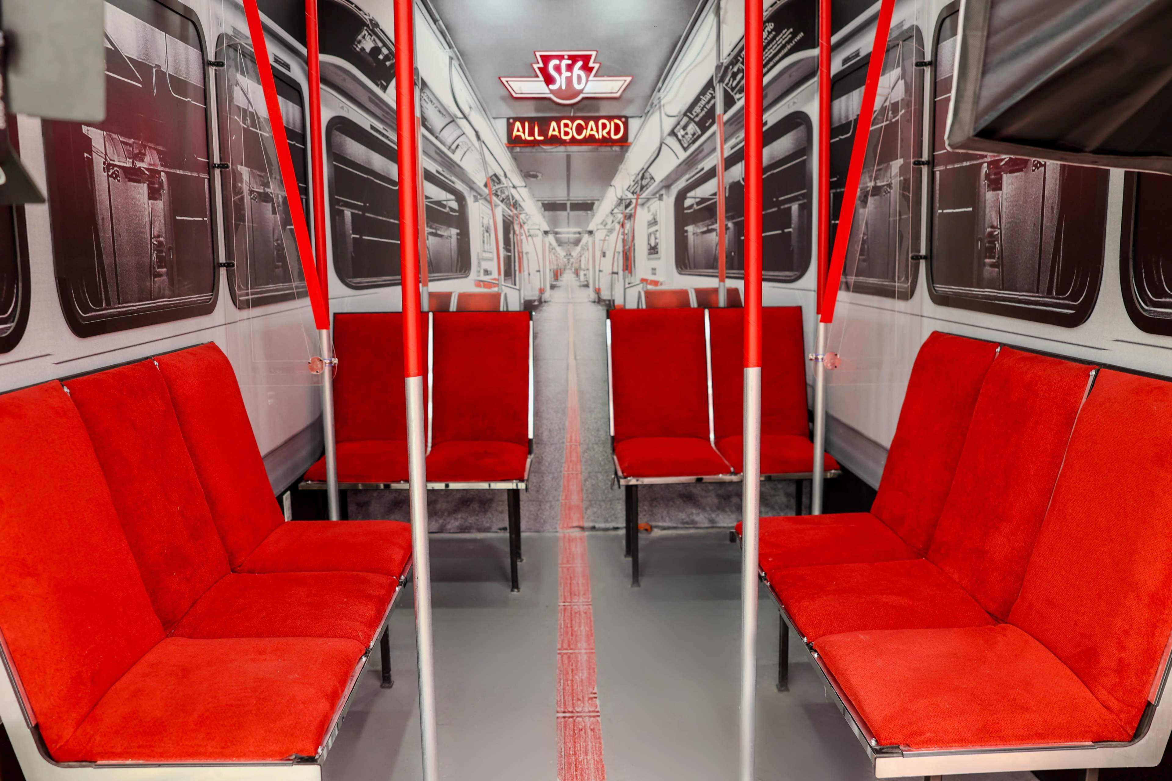 The interior of a subway train features red upholstered seats arranged in pairs with overhead poles and a sign reading "SFG ALL ABCARD."