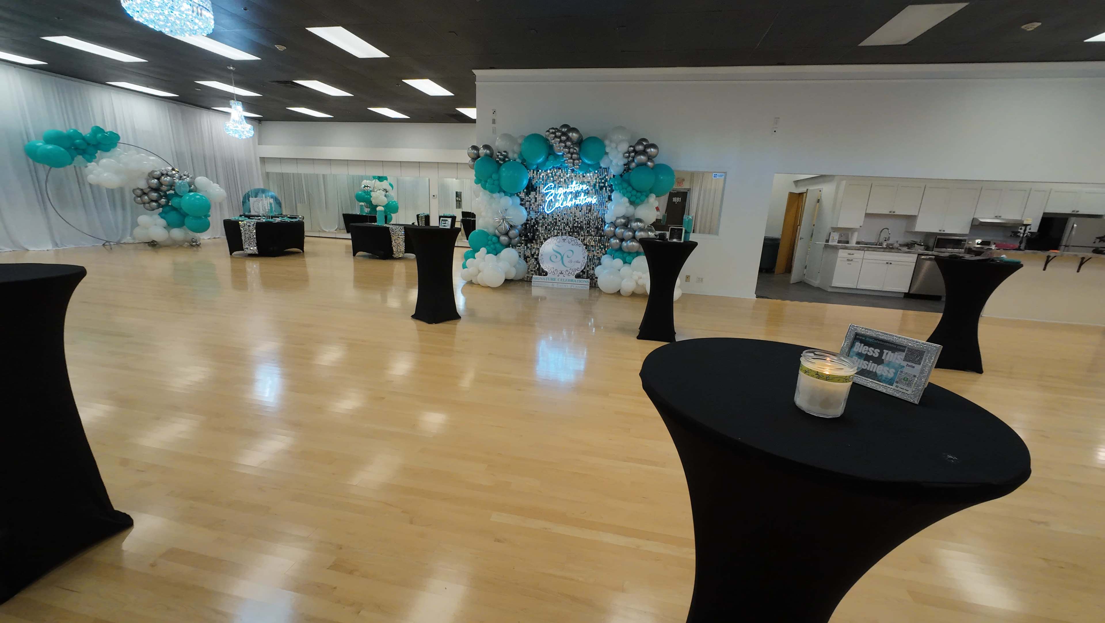 A spacious event venue is decorated with black and teal balloons, matching table covers, and a backdrop featuring a decorative display.