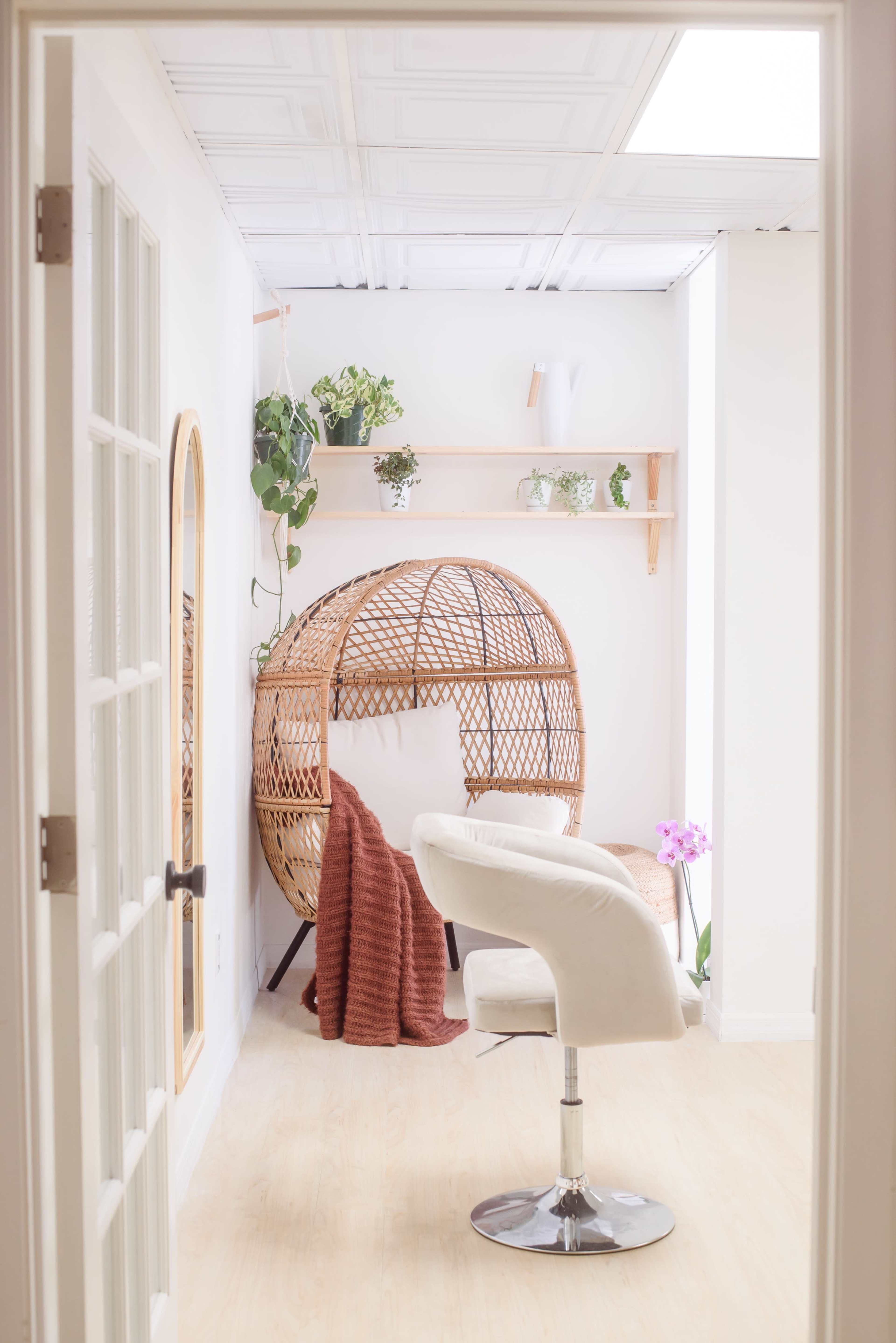 A bright room with a wicker chair, a white stool, and decorative shelves adorned with plants.