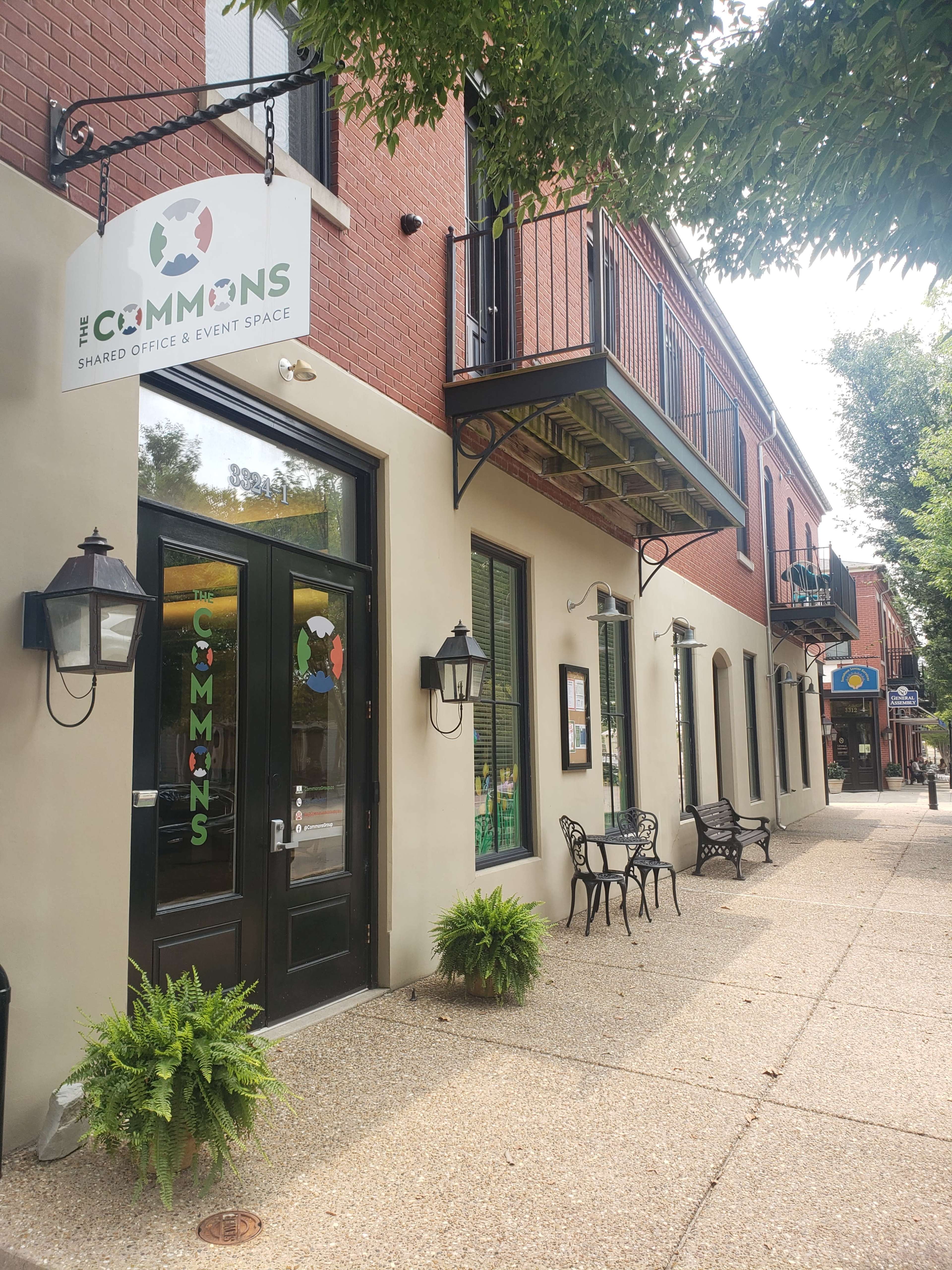 The exterior of a building with a sign for "The Commons," which is a shared office and event space, flanked by benches and decorative plants.