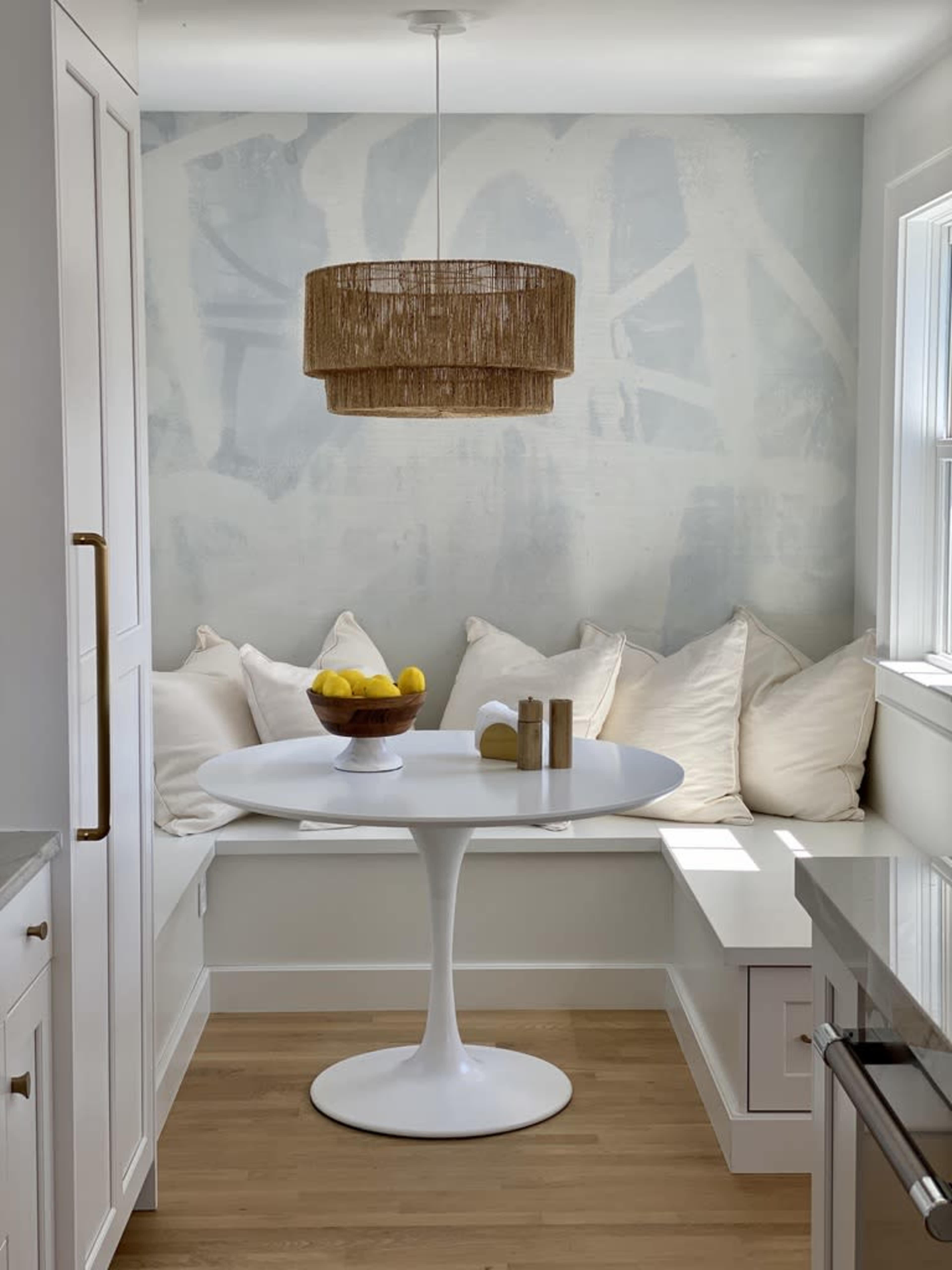 The image shows a cozy breakfast nook featuring a round white table, a bowl of lemons, and a built-in bench with white pillows against a softly painted wall.
