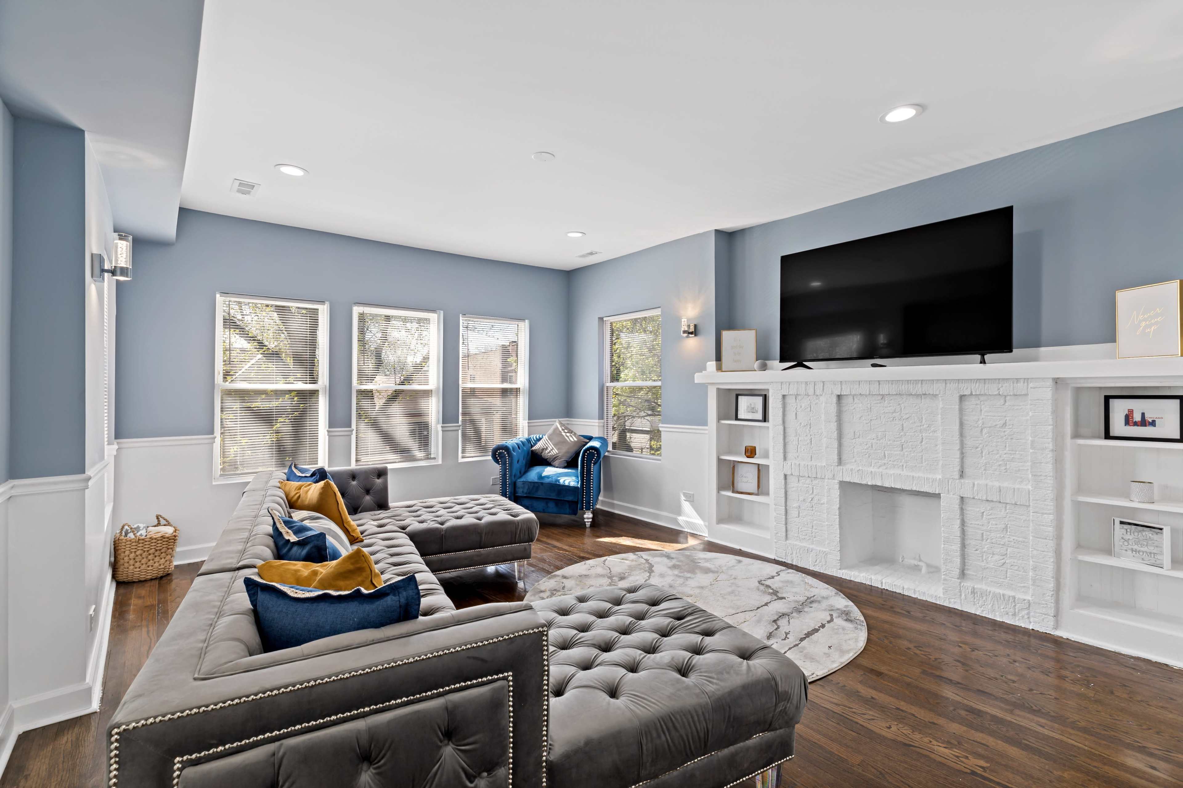 A spacious living room features a tufted sectional sofa, a round area rug, and a large television mounted above a white brick fireplace.