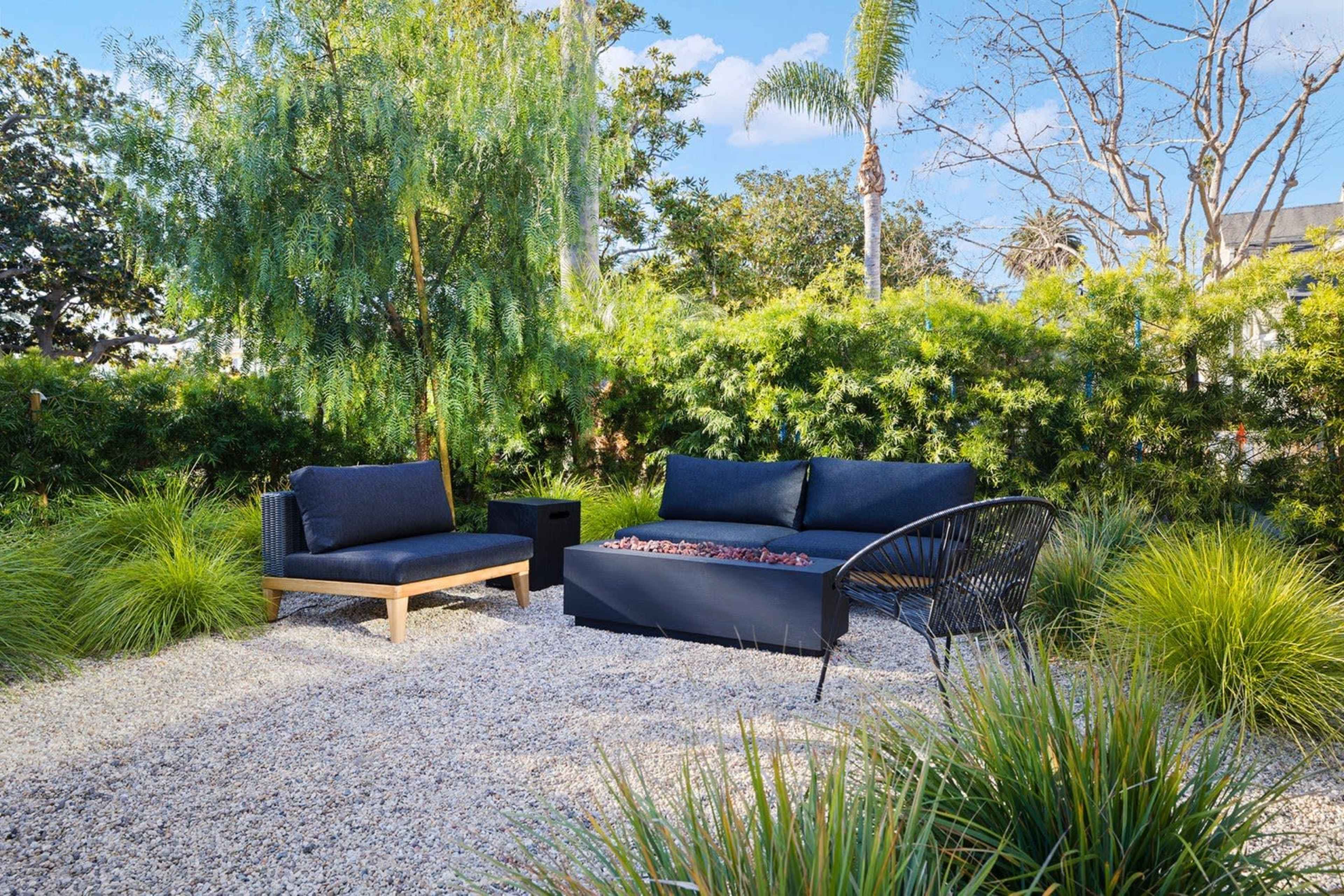 A modern outdoor seating area features a black couch and a matching chair on a gravel surface surrounded by lush greenery and low shrubs.