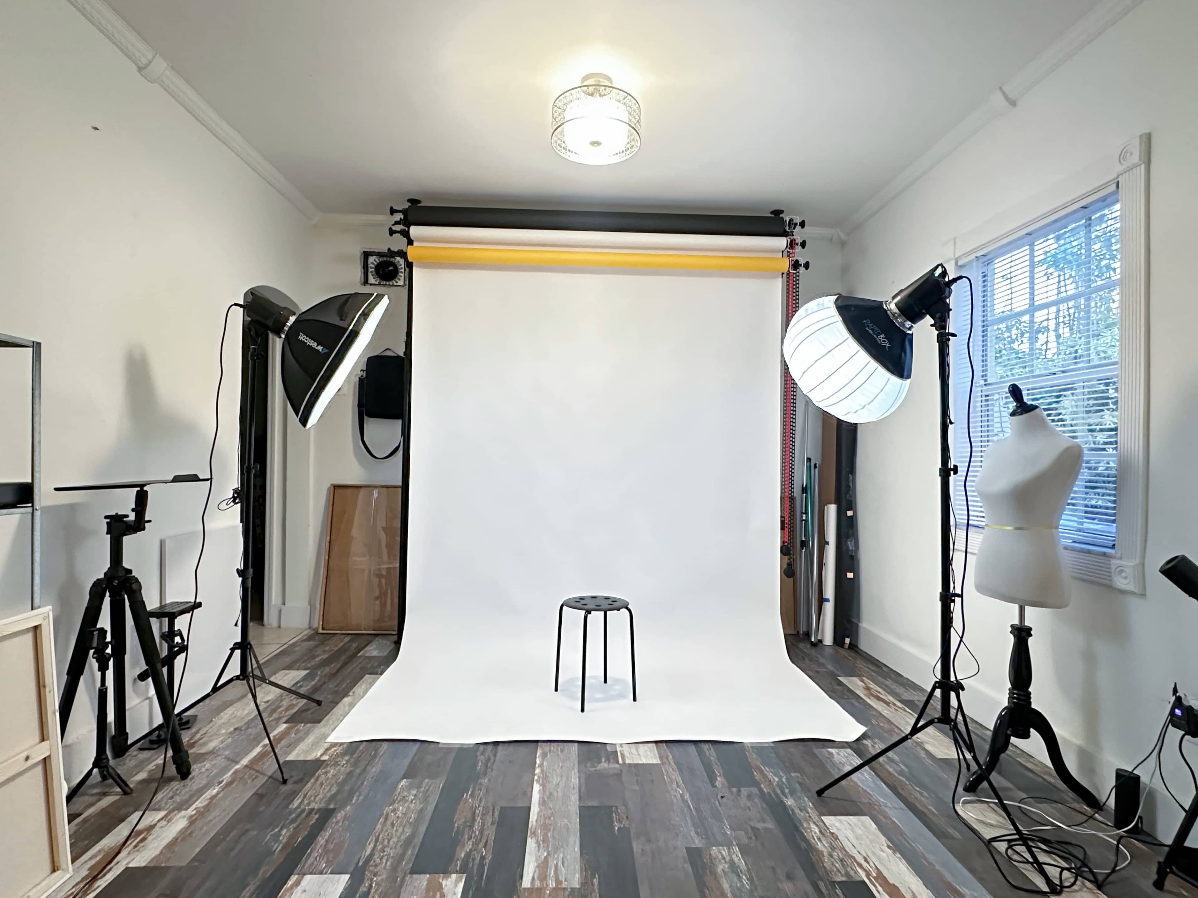 The image shows a photography studio equipped with a white backdrop, a stool at the center, and lighting setup on both sides.