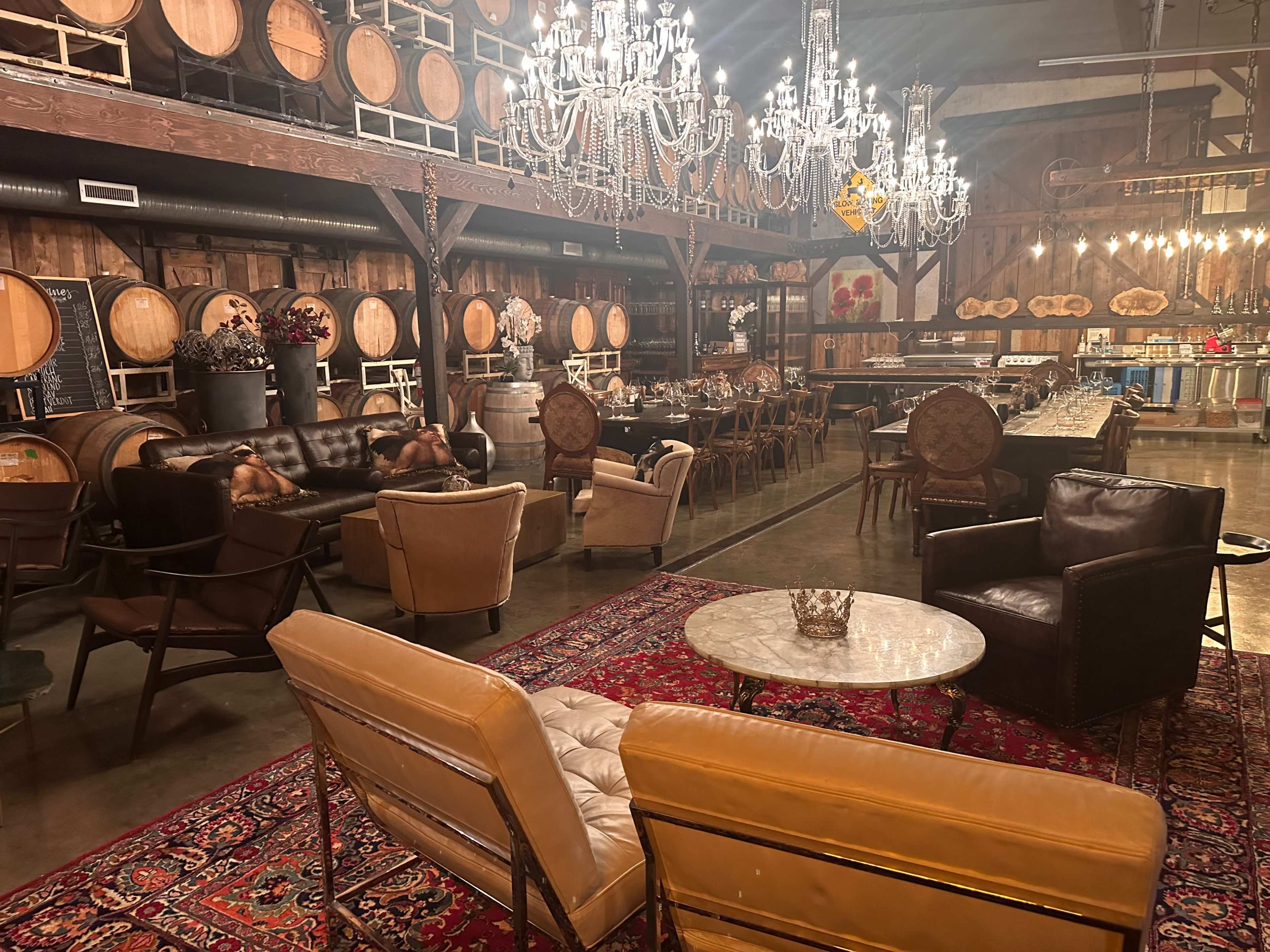 The image shows a rustic winery interior featuring wooden barrels, a chandelier, and seating arrangements on a colorful rug.