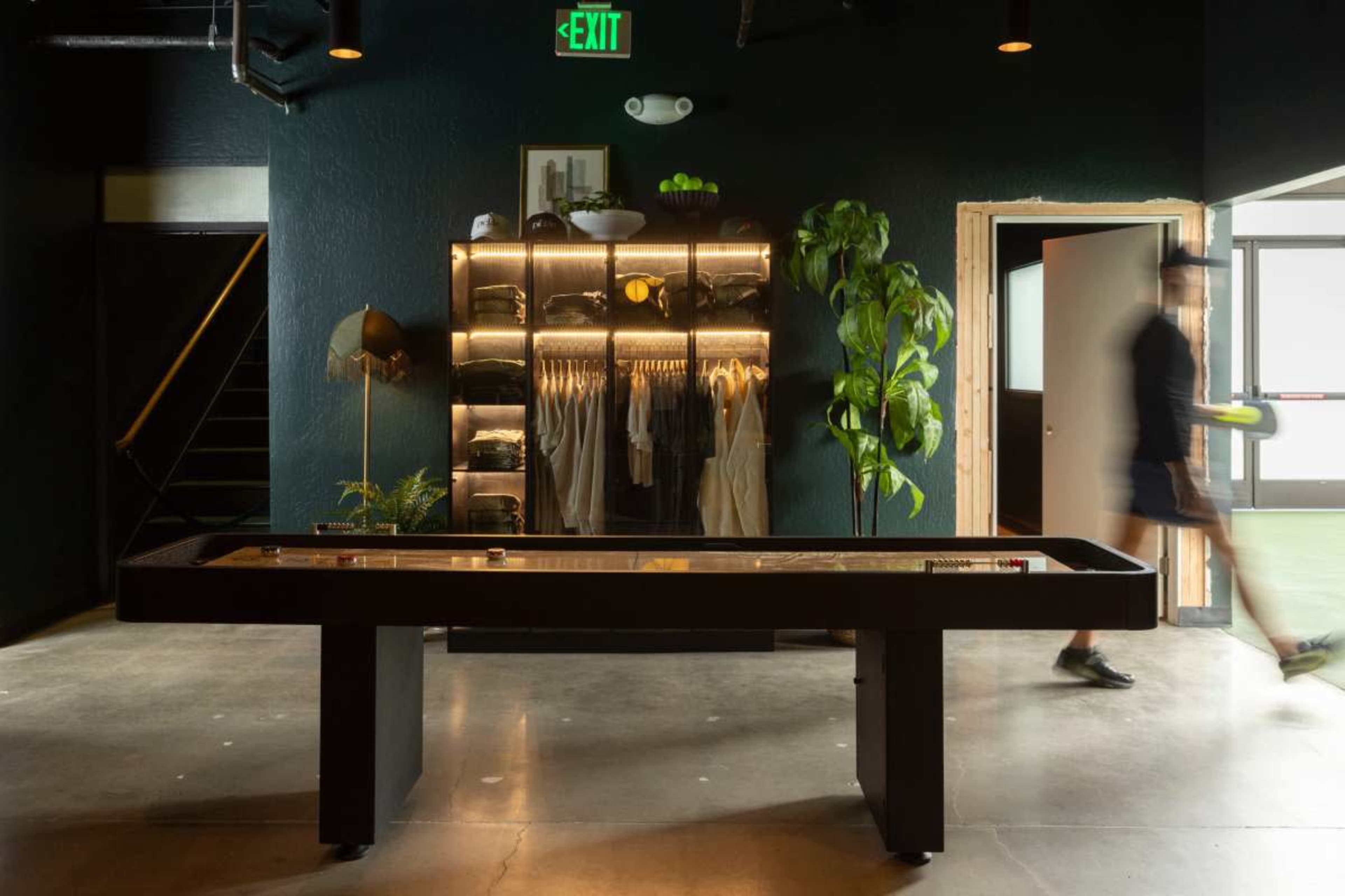 The image shows a modern interior featuring a shuffleboard table, a clothing display with hangers, a potted plant, and a person walking past a doorway.