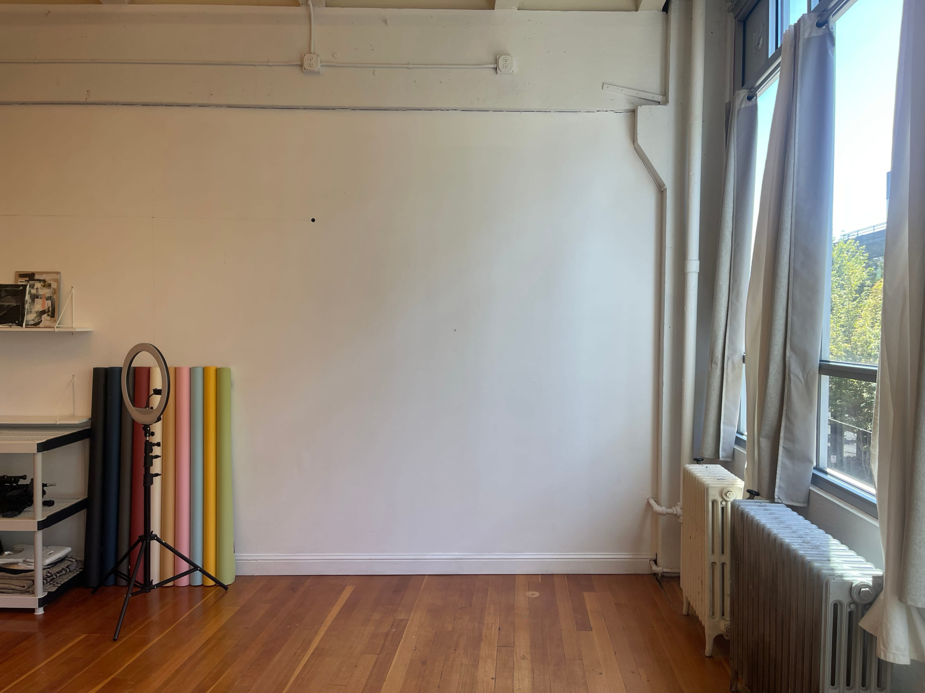 An empty room with hardwood floors, a large window, a mirror on a stand, and vertical color swatches leaning against the wall.