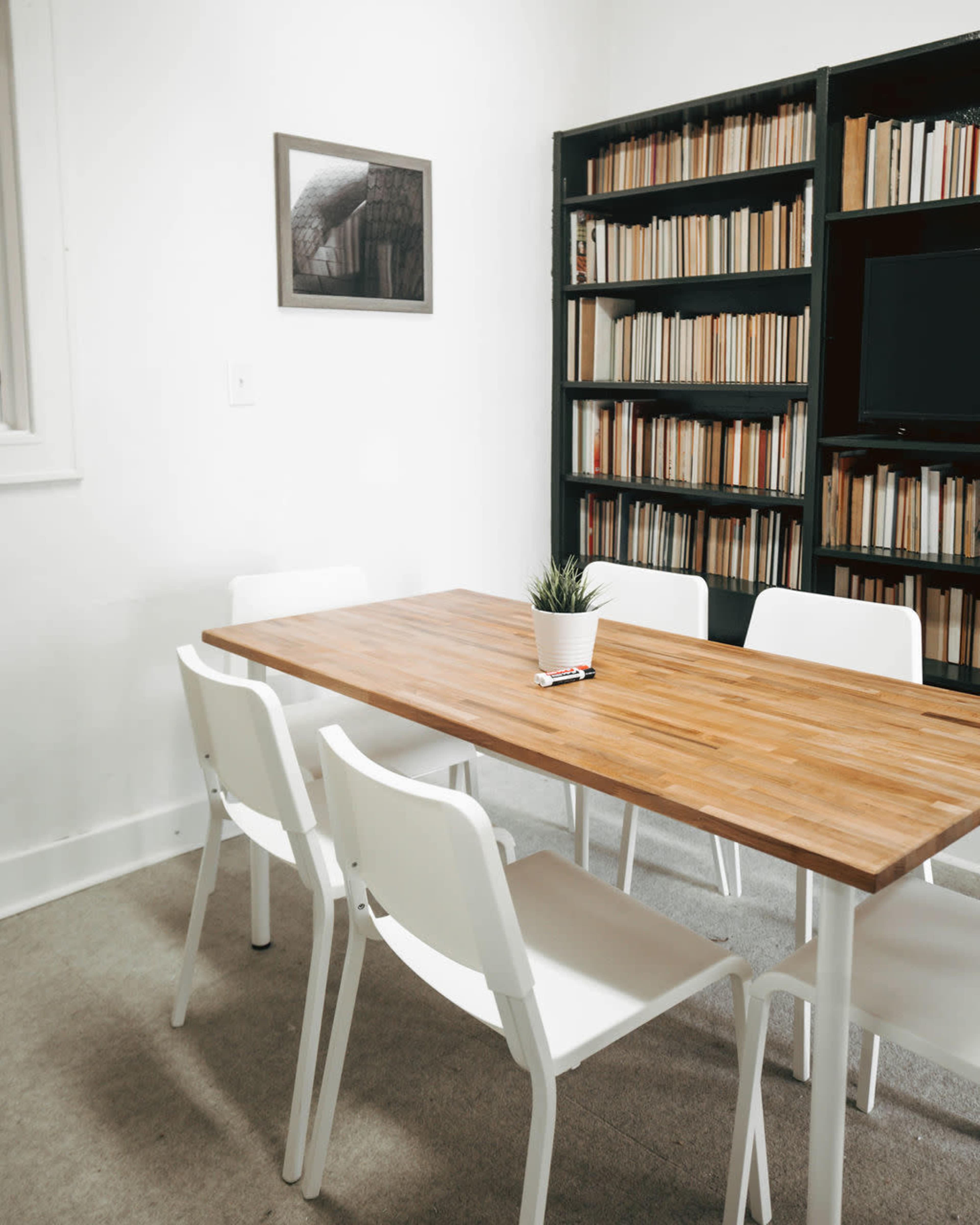 A wooden table is center in a bright room surrounded by four white chairs, with a bookshelf filled with books and a small potted plant on the table.