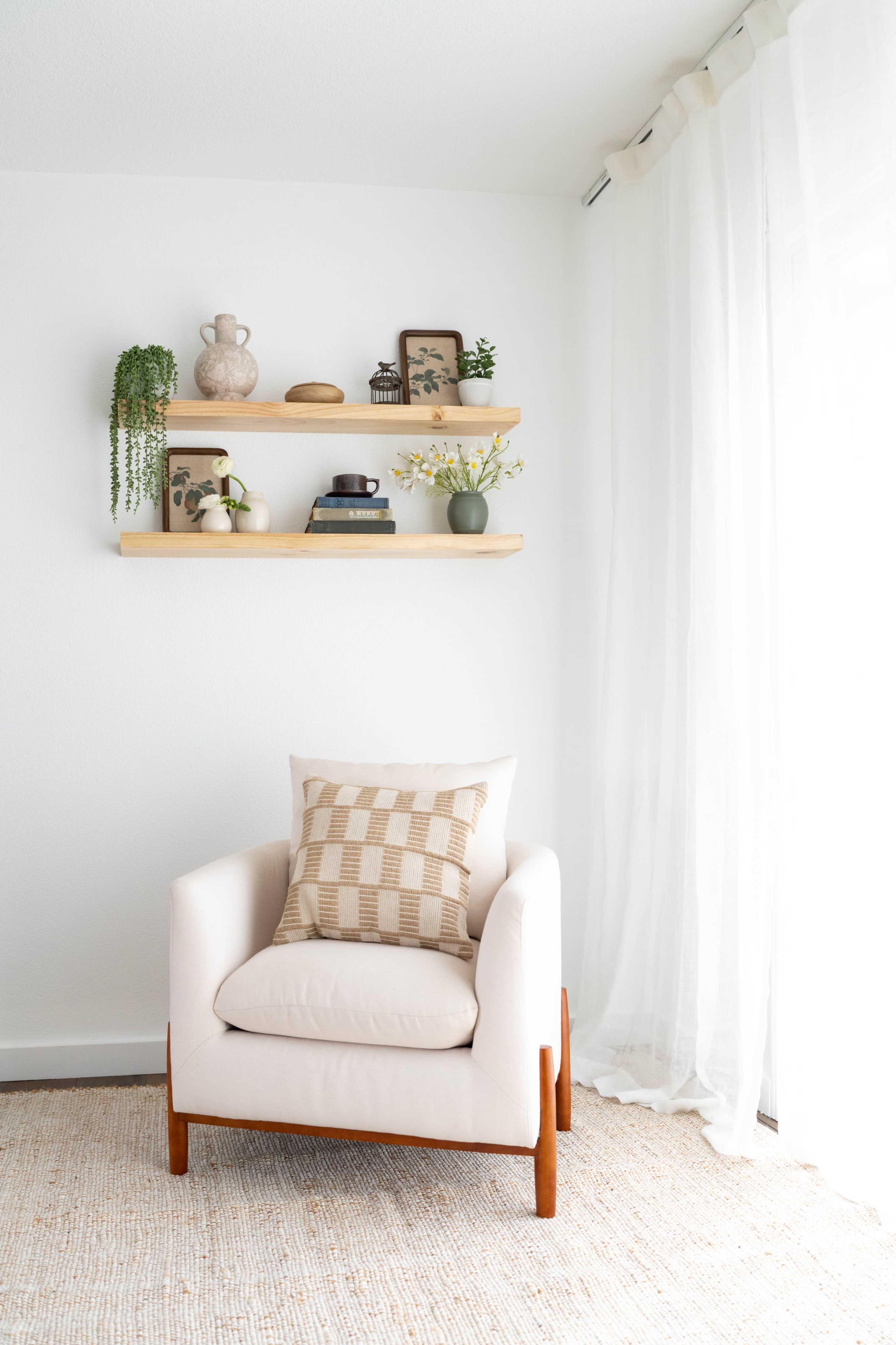 The image shows a minimalist living space featuring a white armchair with a patterned pillow, a light-colored rug, and a wooden shelf above that holds decorative items and plants, complemented by sheer curtains.