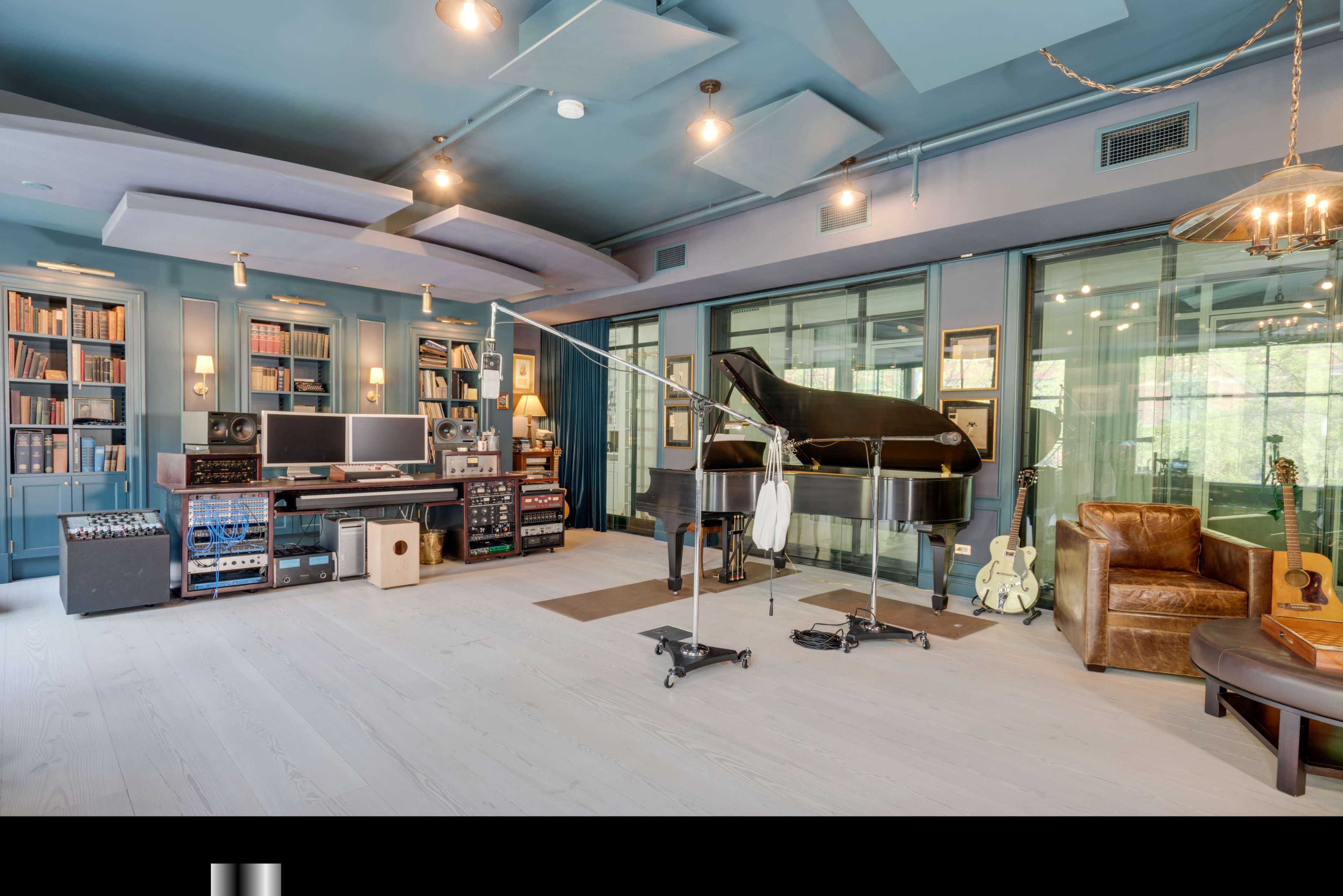 A well-equipped music studio featuring a grand piano, audio mixing equipment, and guitars, with large windows allowing natural light.