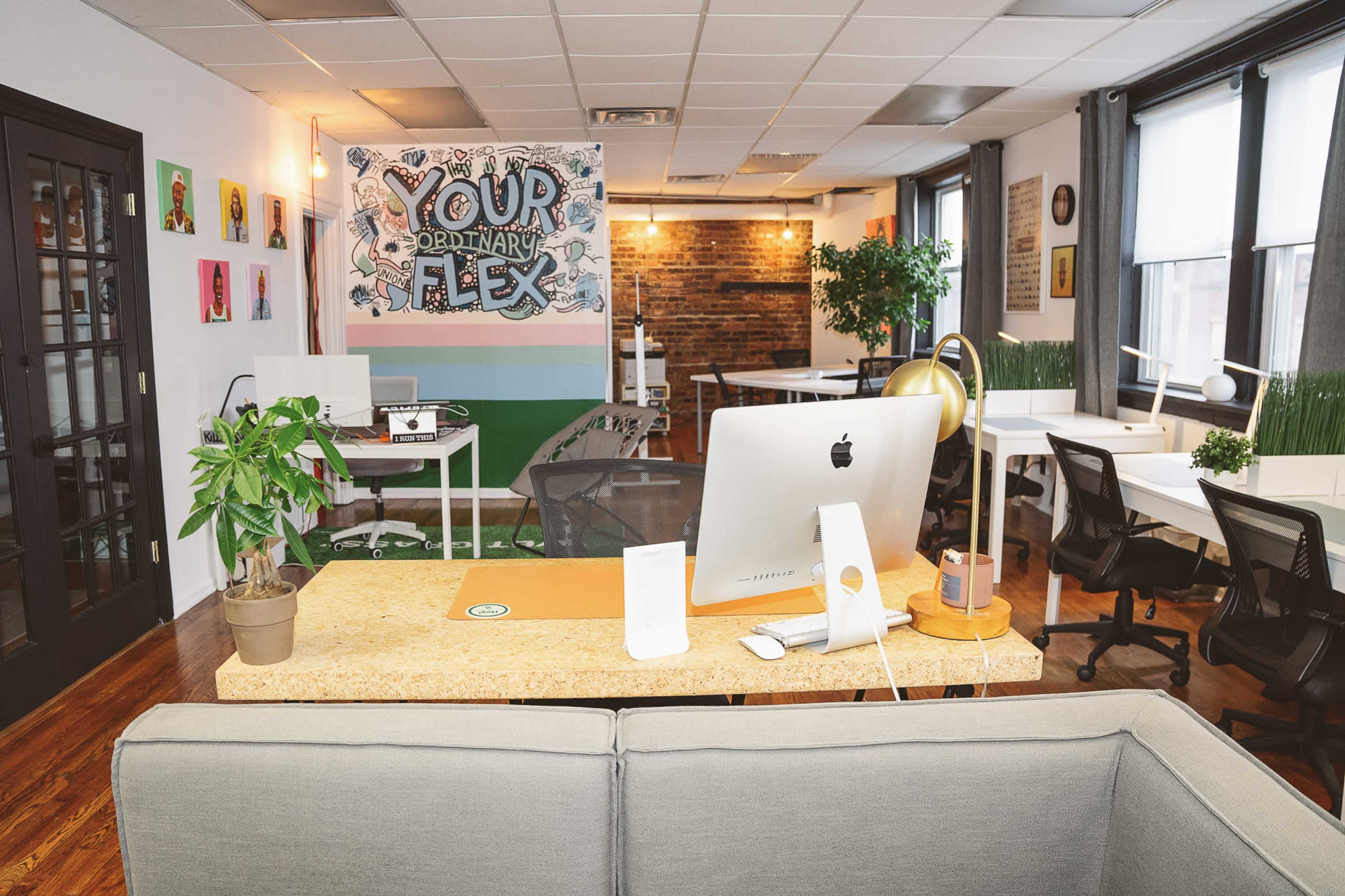A modern office space with multiple workstations, a decorative mural on the wall, plants, and a large desk with an Apple computer.
