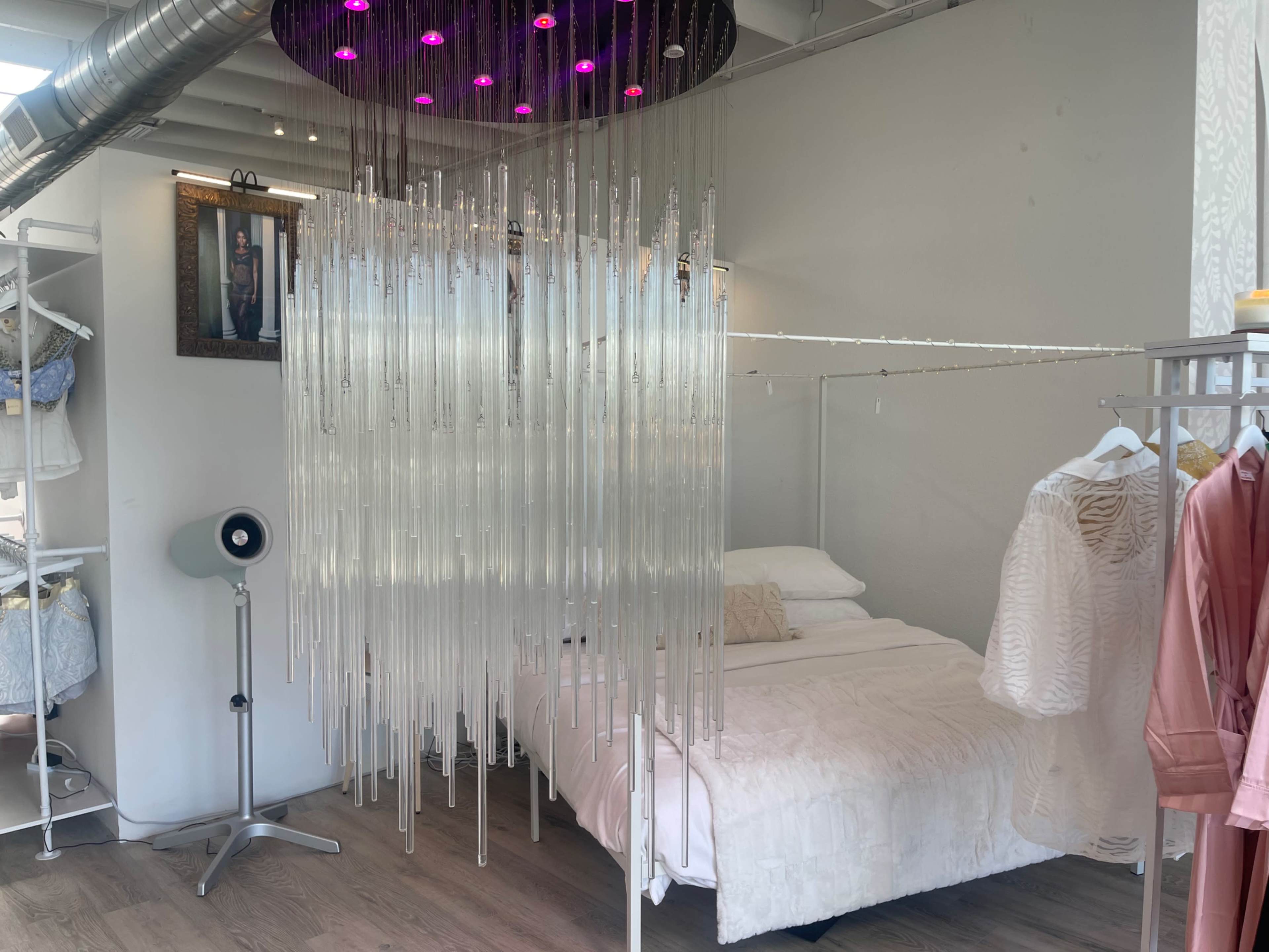 A modern bedroom area features a bed with white linens, separated from a clothing display by a hanging installation of clear tubes, beneath a circular ceiling light with purple accents.