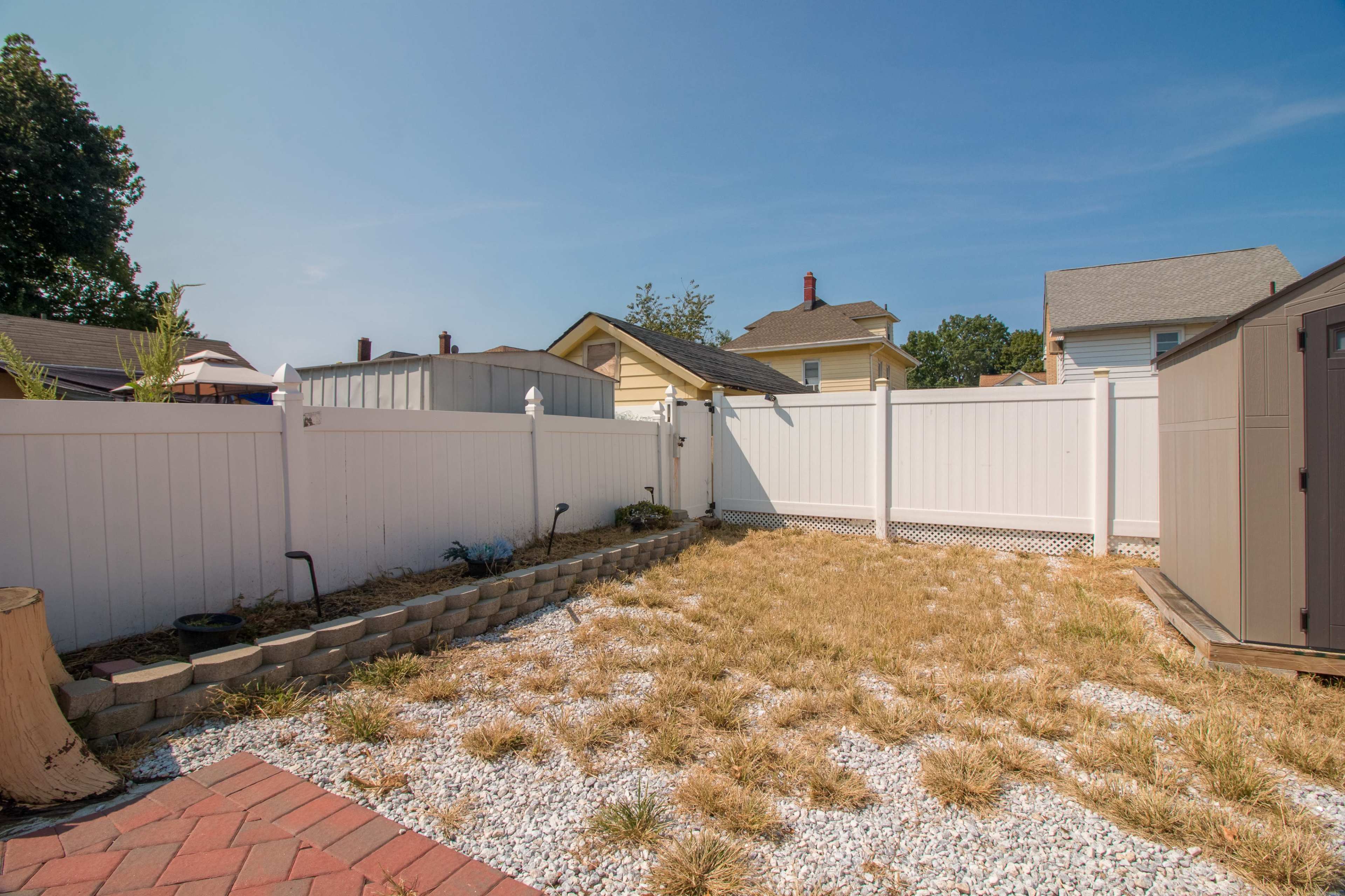 The scene shows a small, fenced backyard with dry grass, gravel, and a storage shed.