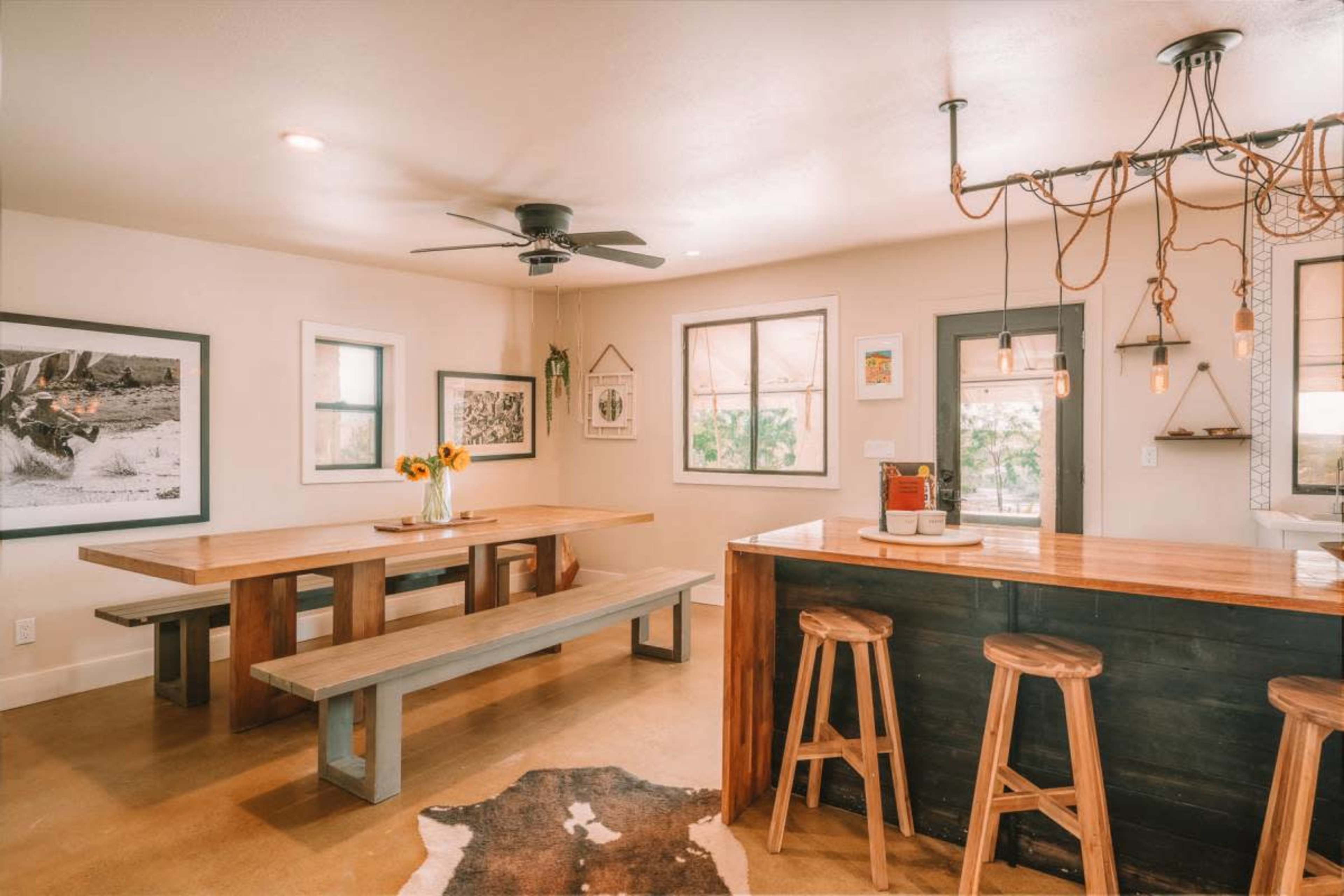 The image depicts a modern dining area featuring a large wooden table with benches, a bar counter with stools, and light fixtures hanging overhead.