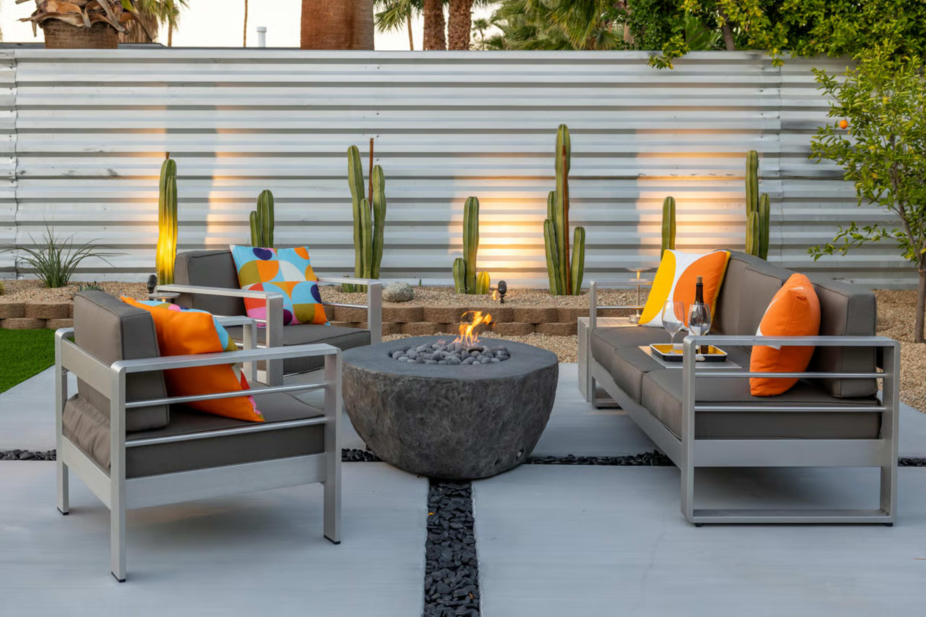 A modern outdoor seating area features gray couches with colorful pillows arranged around a circular stone fire pit, surrounded by cacti and illuminated by soft lighting.