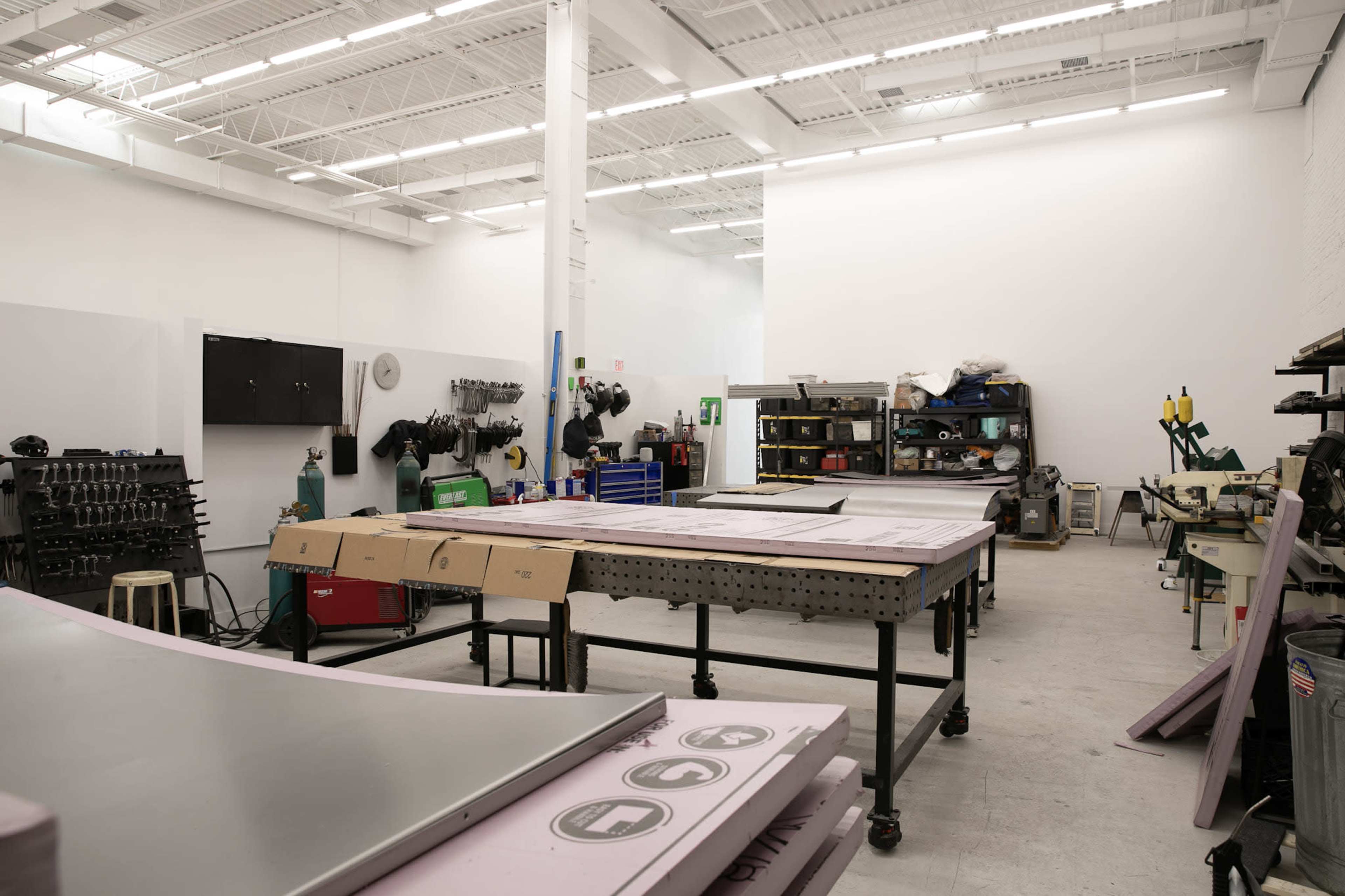 The image shows a workshop with various tools and equipment arranged on worktables and shelves, featuring a large, open space with a concrete floor.