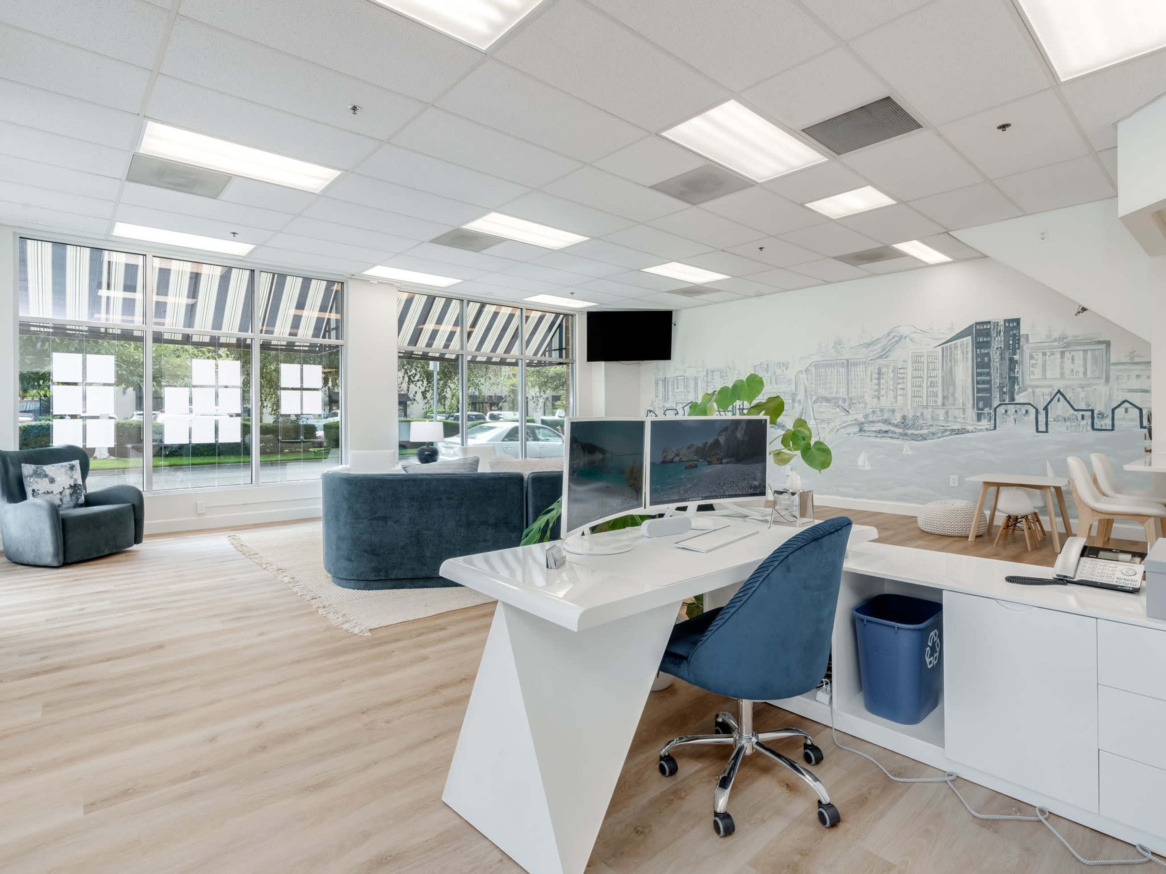The image shows a modern office space featuring a reception desk, a blue couch, a dining area, and large windows with a mural on the wall.