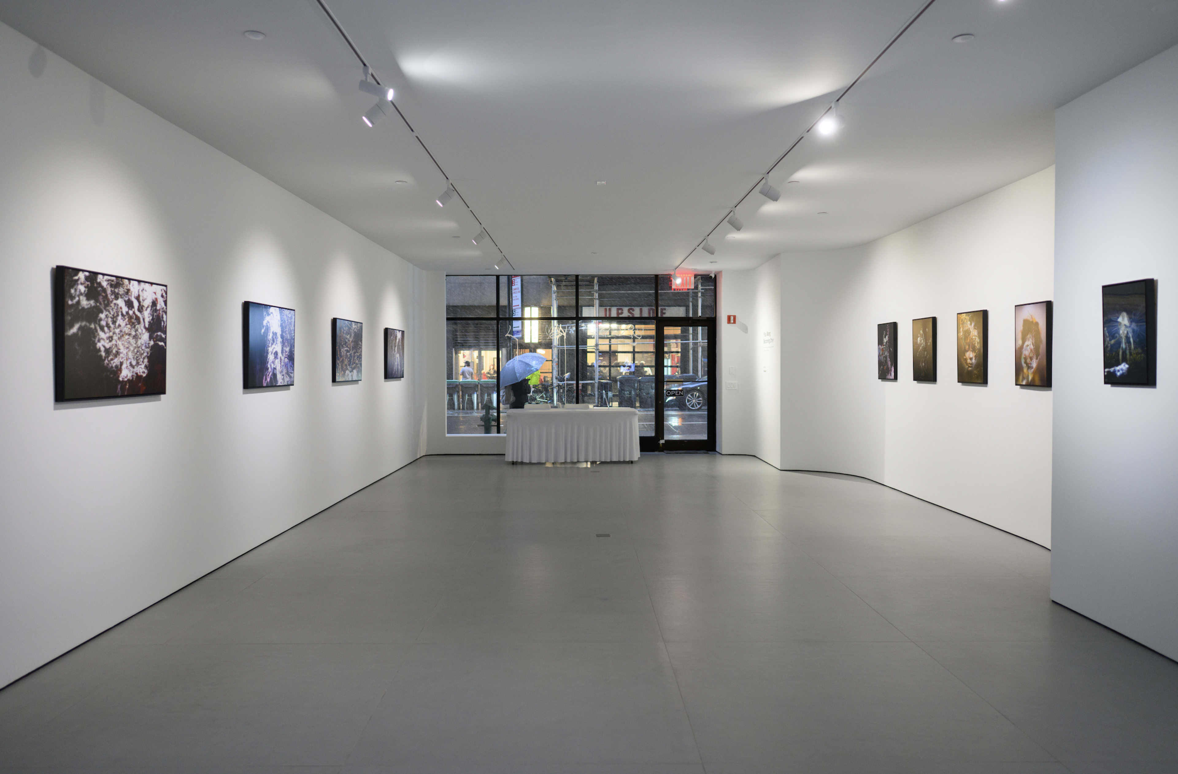 The image shows an art gallery with white walls displaying framed photographs, and a large window revealing an outdoor street with a person holding an umbrella.