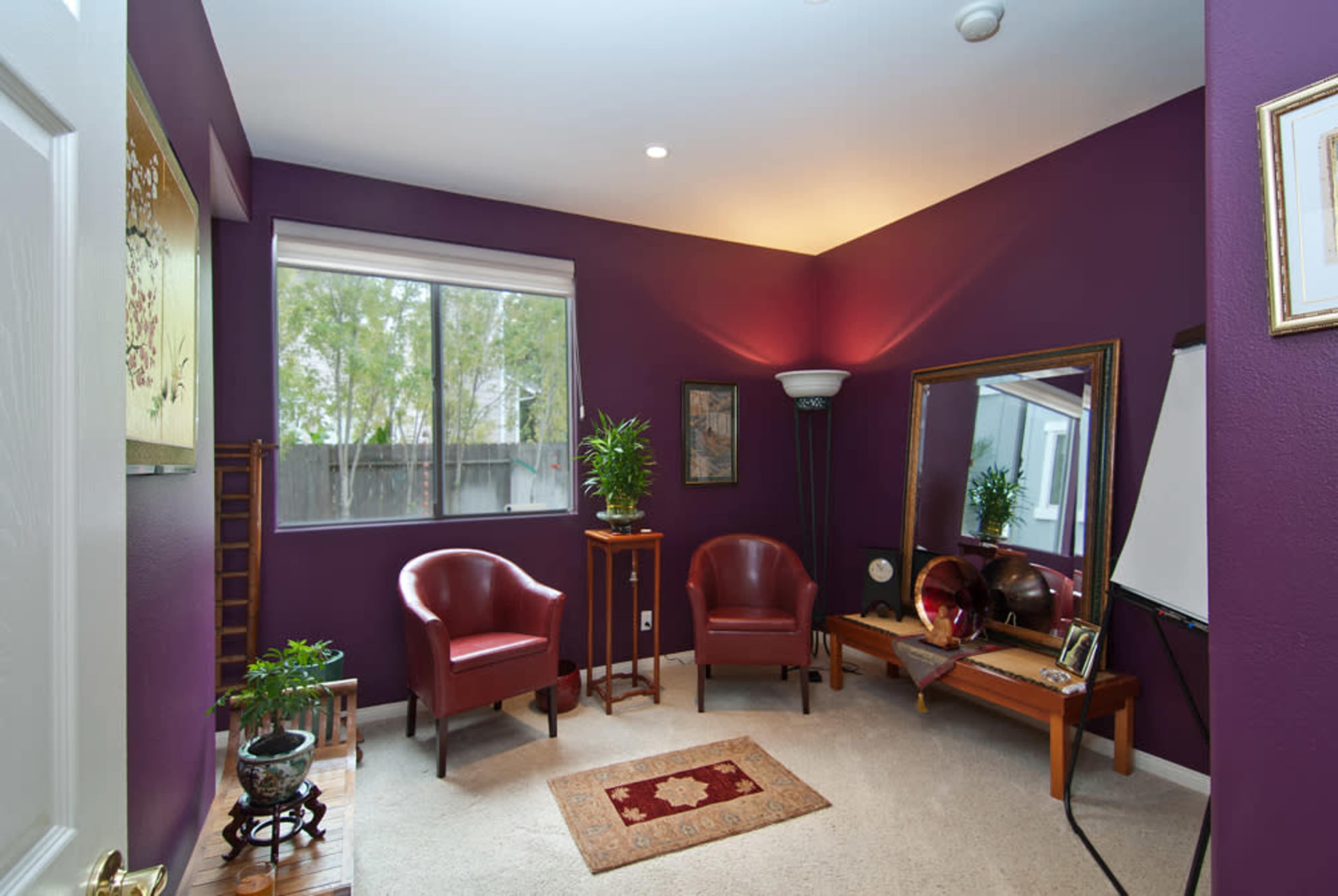 A small, cozy room with purple walls, two red chairs, a mirror, a table with plants, and a window letting in natural light.