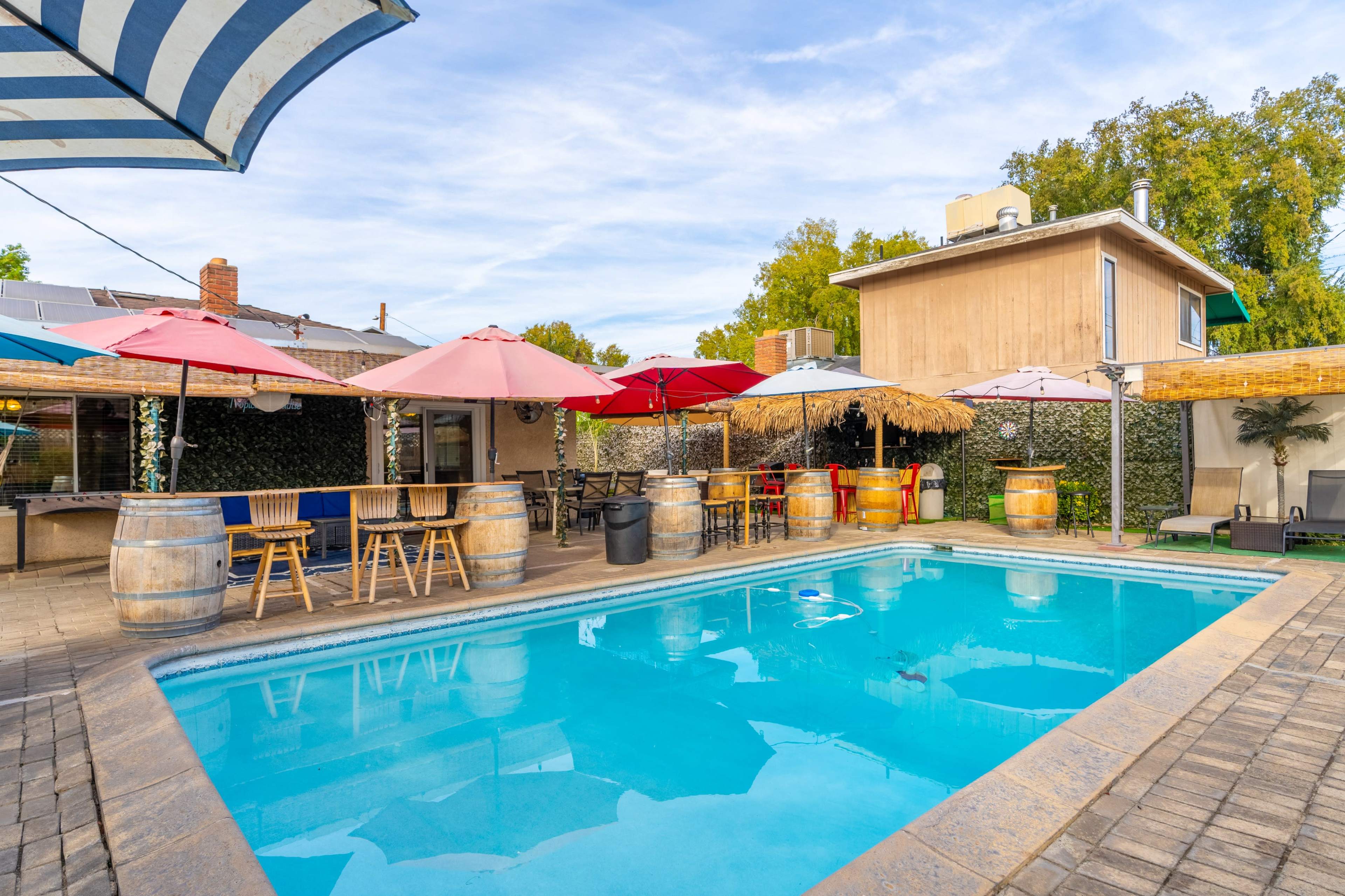 A pool area surrounded by colorful umbrellas, wooden barrels, and lounge chairs, with a house in the background.