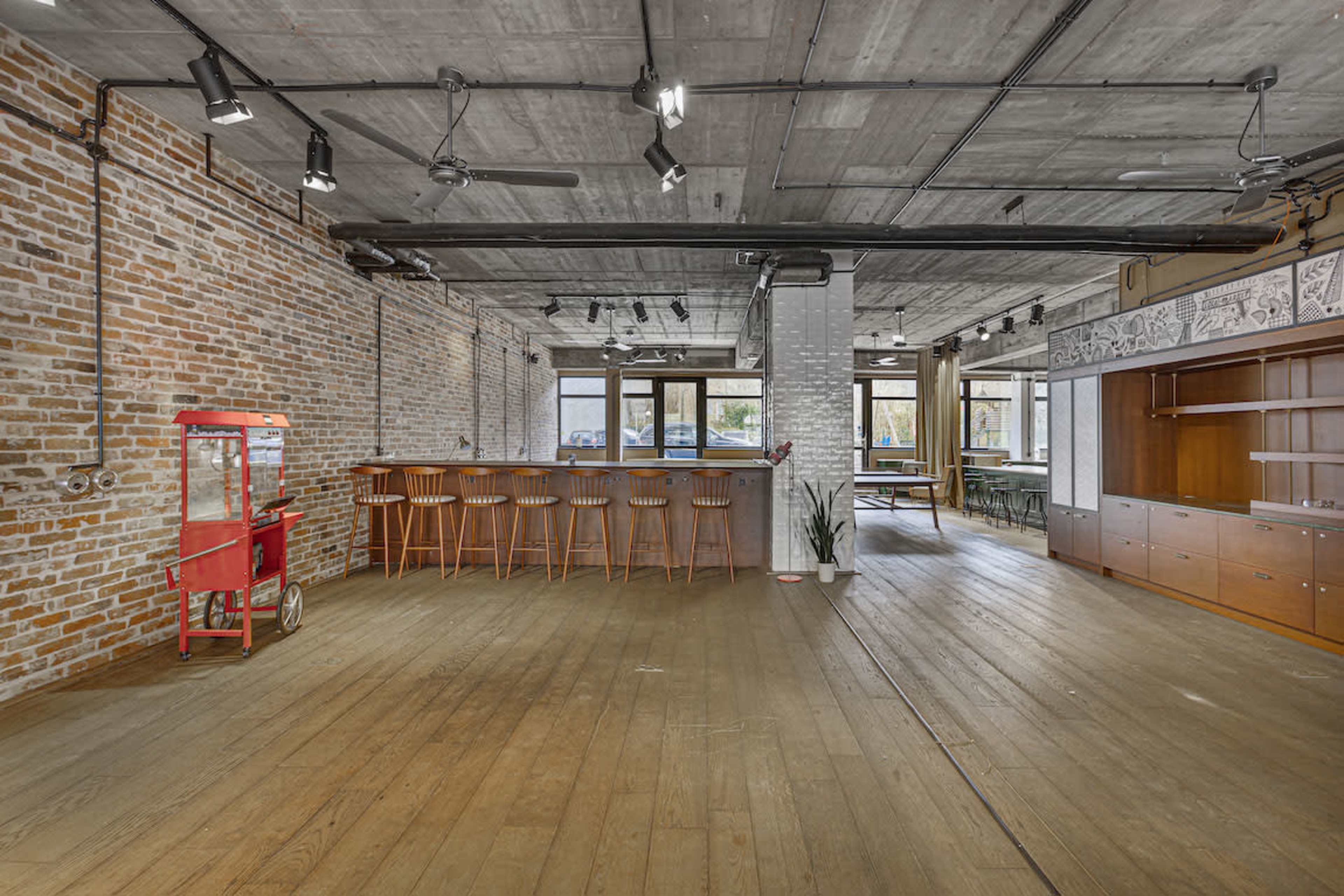 An open industrial-style interior space featuring exposed brick walls, wooden flooring, a bar area with stools, and a red cart in the corner.