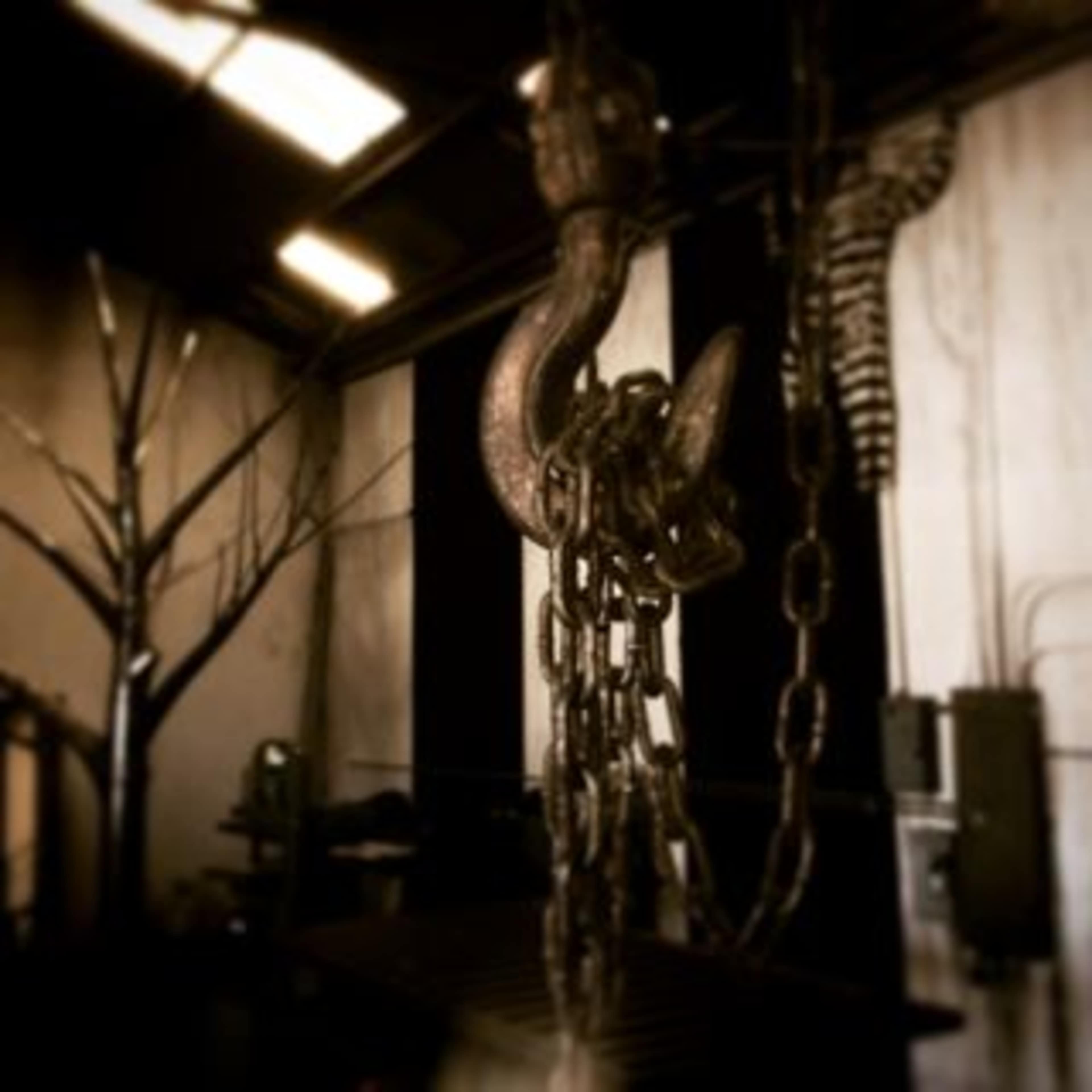 A large metallic hook with chains hangs in a dimly lit industrial space featuring stark walls and a barren tree in the background.