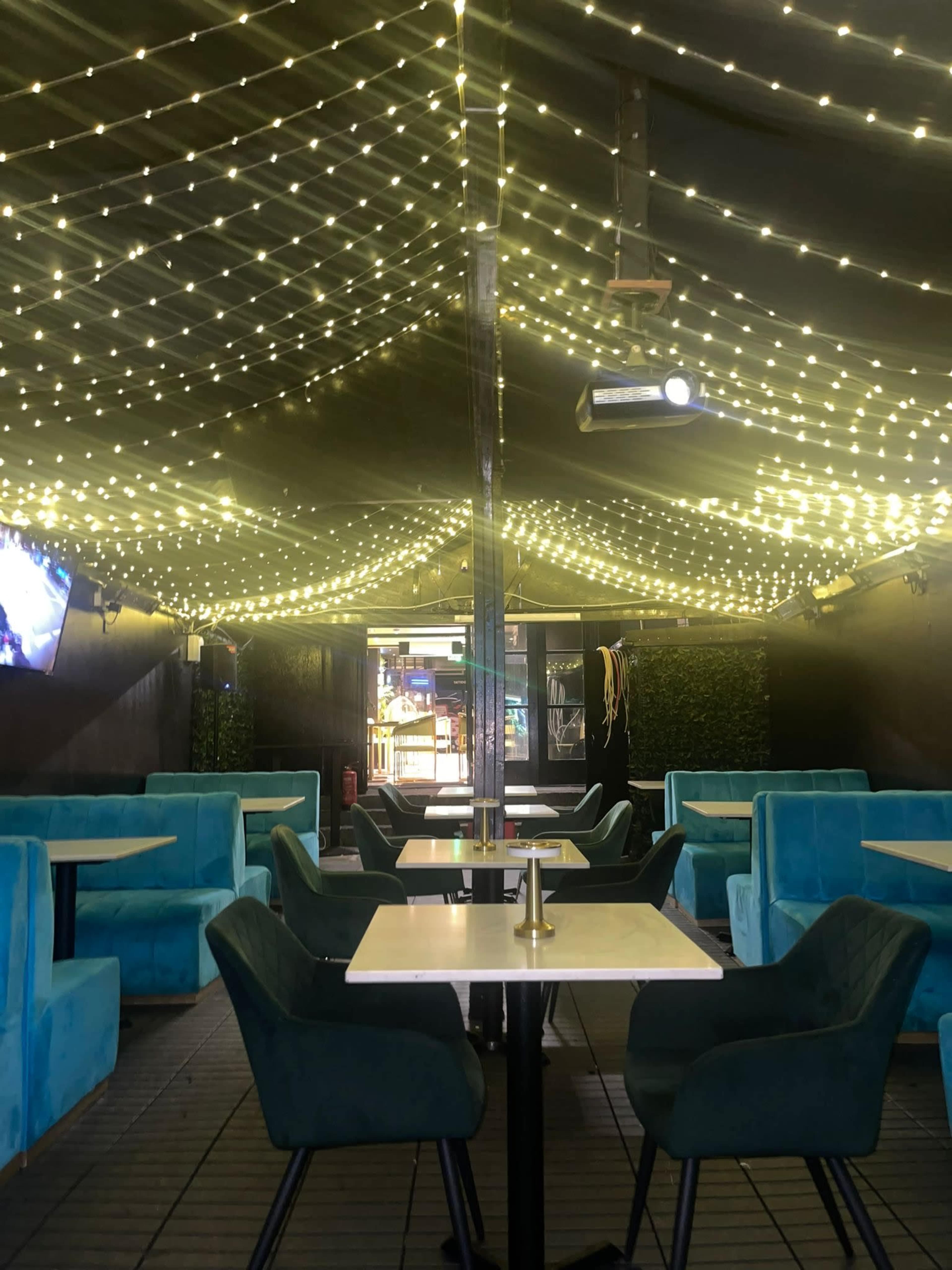 The image shows an outdoor dining area with teal seating and string lights illuminating the ceiling.