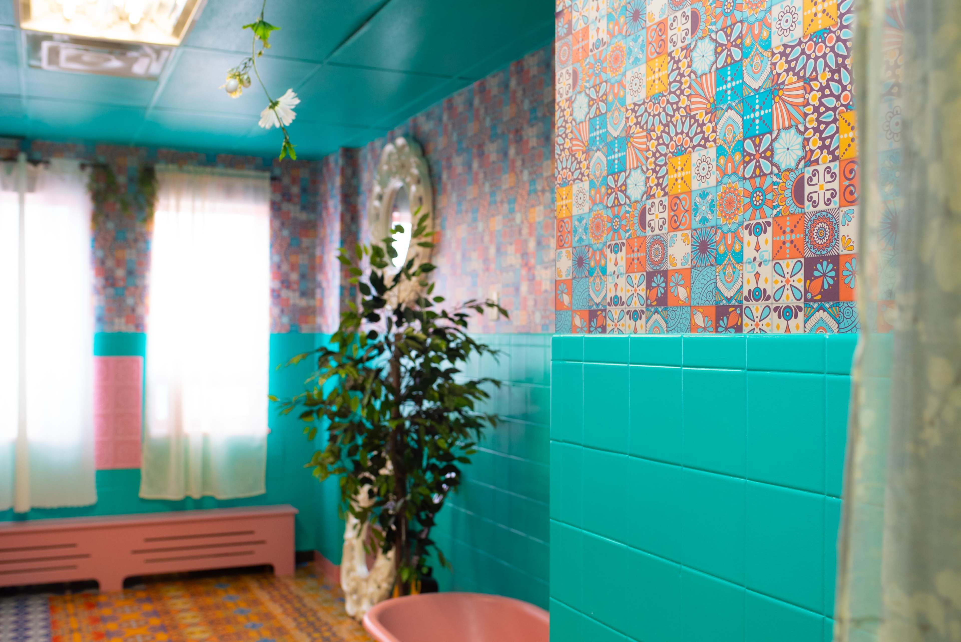 A brightly colored bathroom features patterned tile walls, a pink bathtub, and a plant in the corner, with sunlight streaming through sheer curtains.