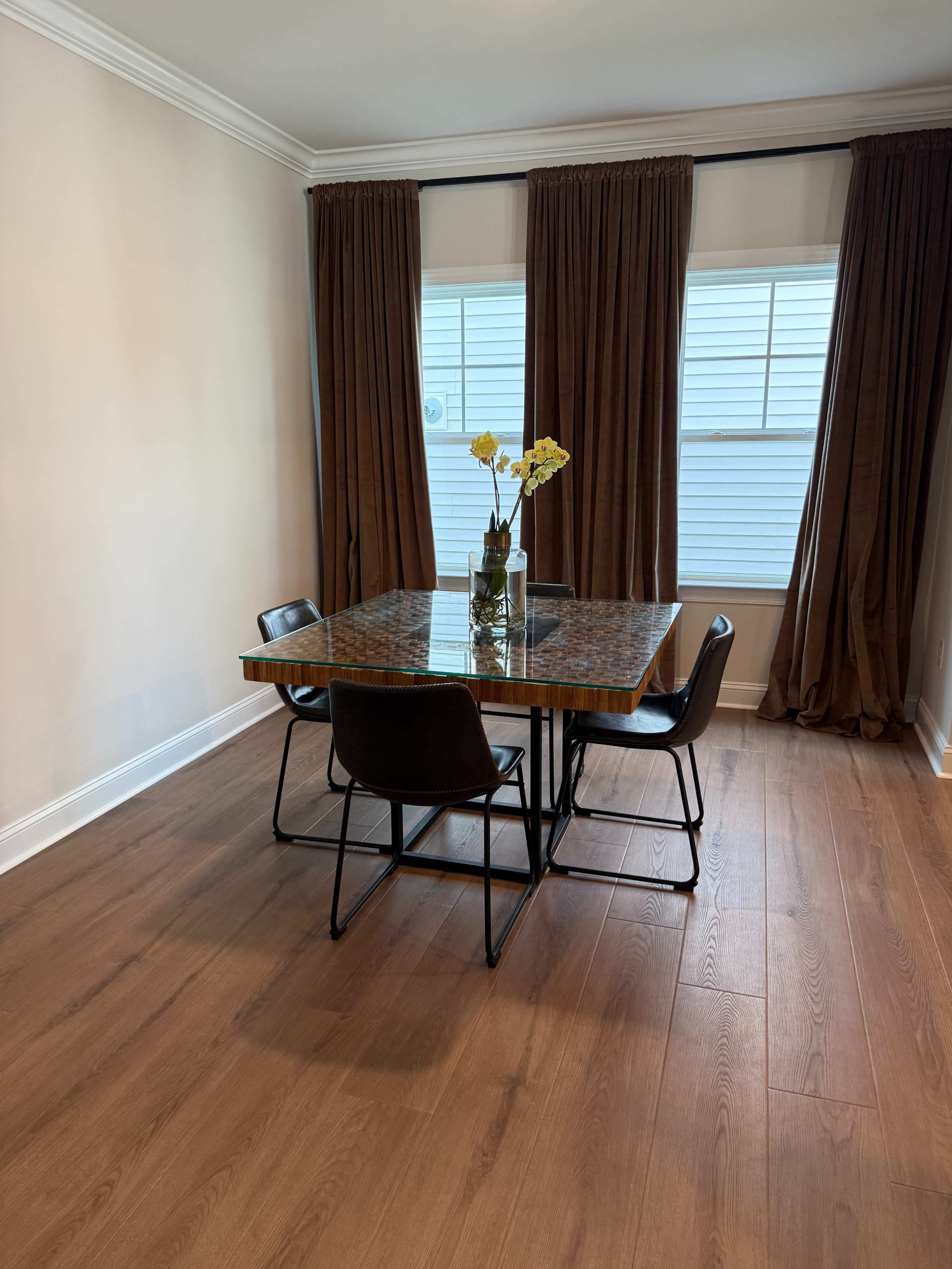 A square dining table with a glass top and four black chairs is positioned in a room with two tall windows covered by brown curtains.