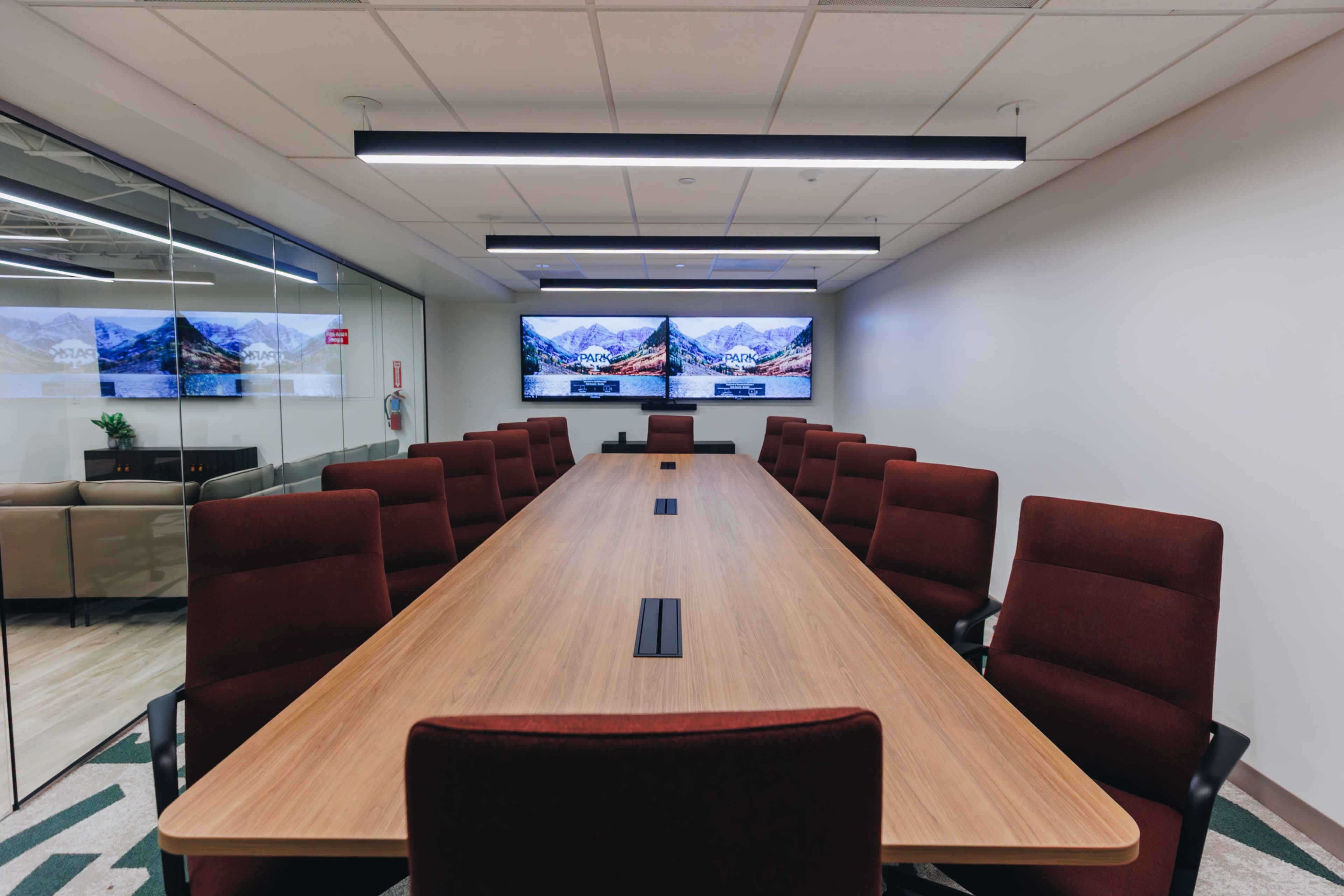 A long wooden conference table with burgundy chairs is set up in a modern meeting room that features two large screens displaying mountain landscapes on the walls.
