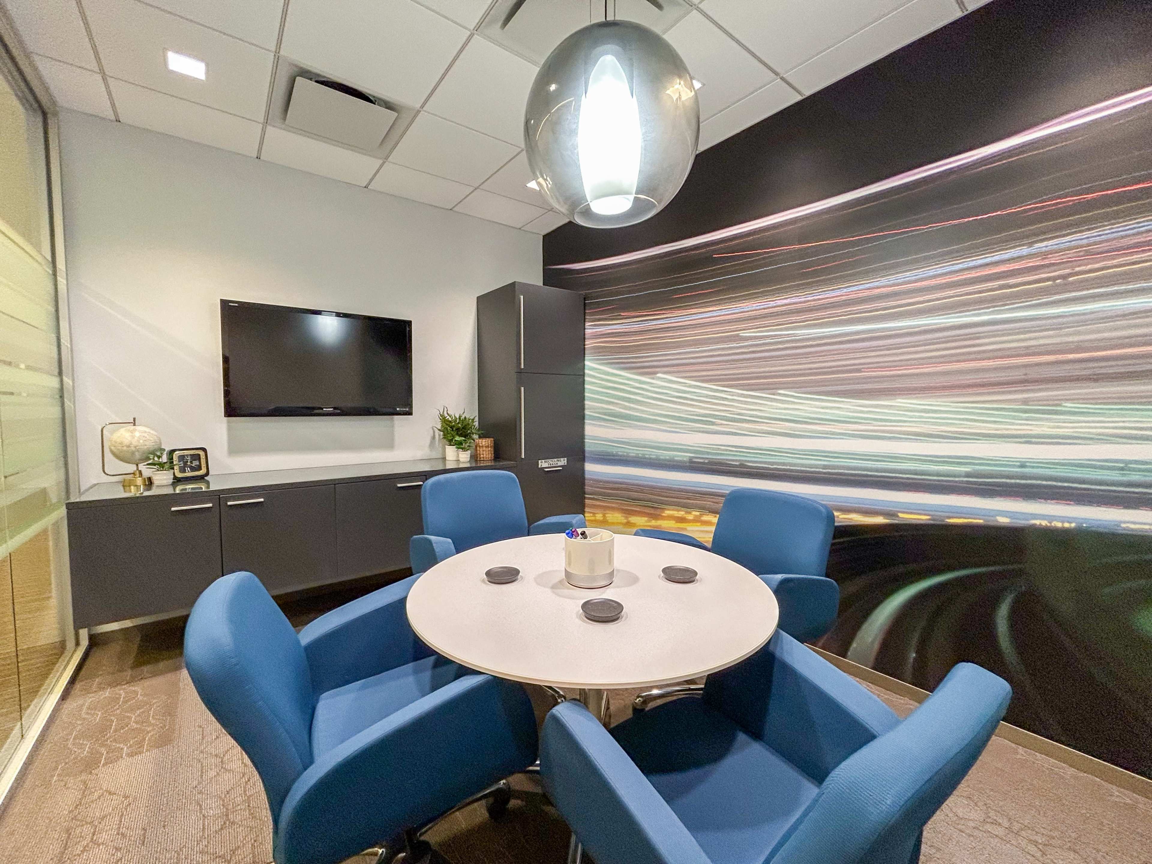 The image shows a modern conference room with a circular table surrounded by four blue chairs, a television mounted on the wall, and a vibrant, abstract mural.
