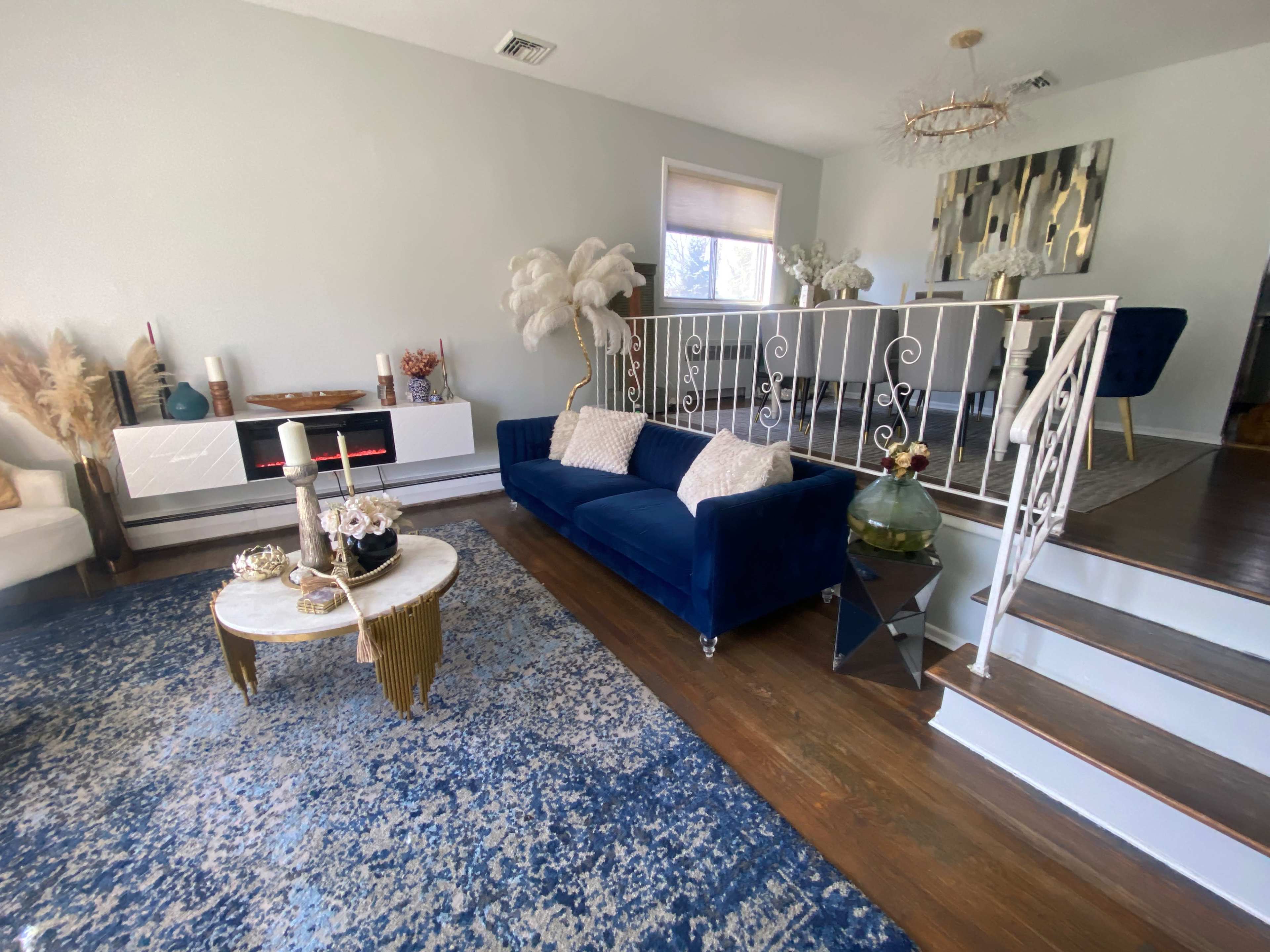 The living room features a blue sofa, a round coffee table, and a staircase leading to a dining area with modern decor.