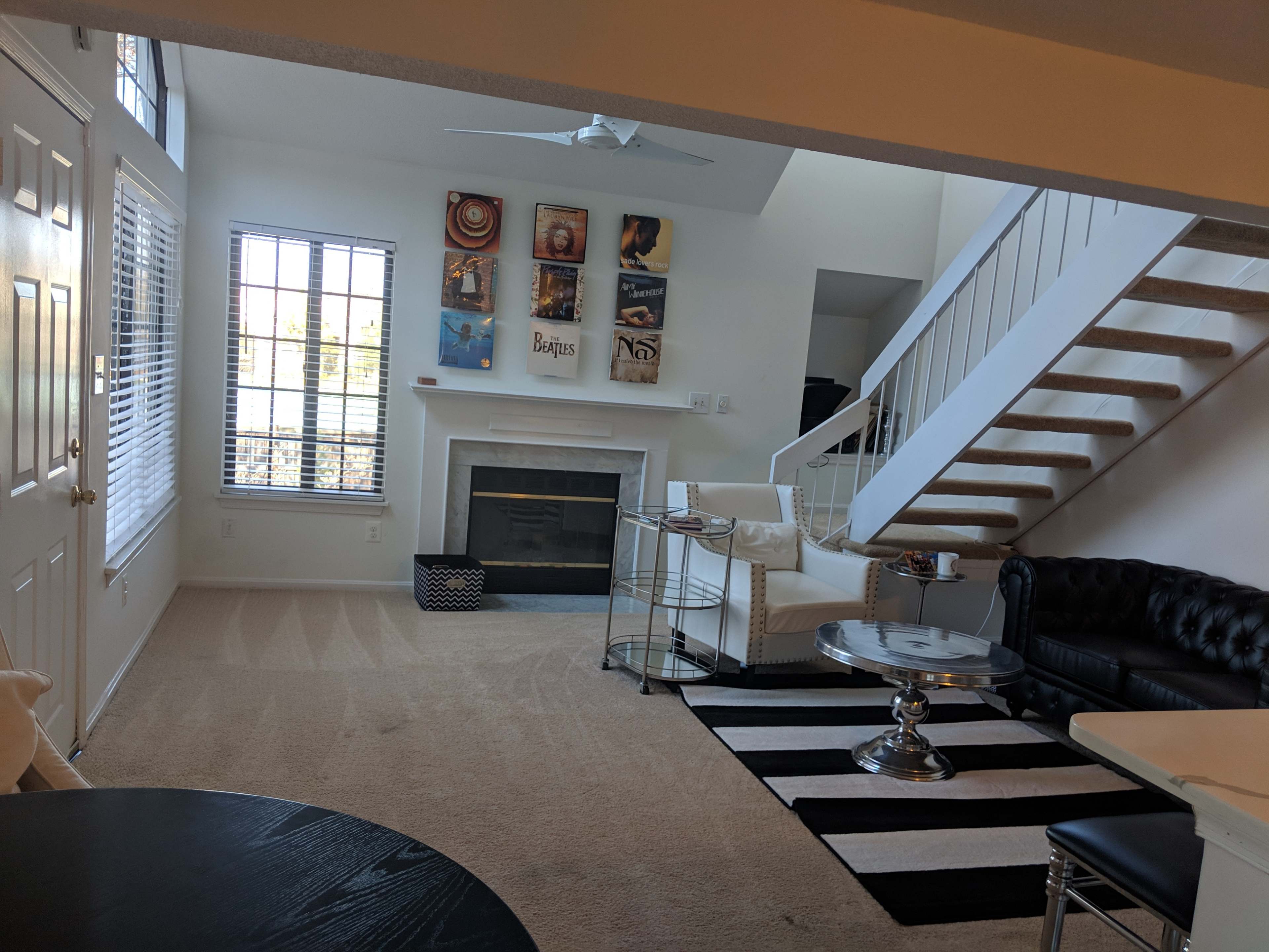 A living room with a fireplace, a black leather couch, a white sofa, a staircase, and a large window allowing sunlight to enter.