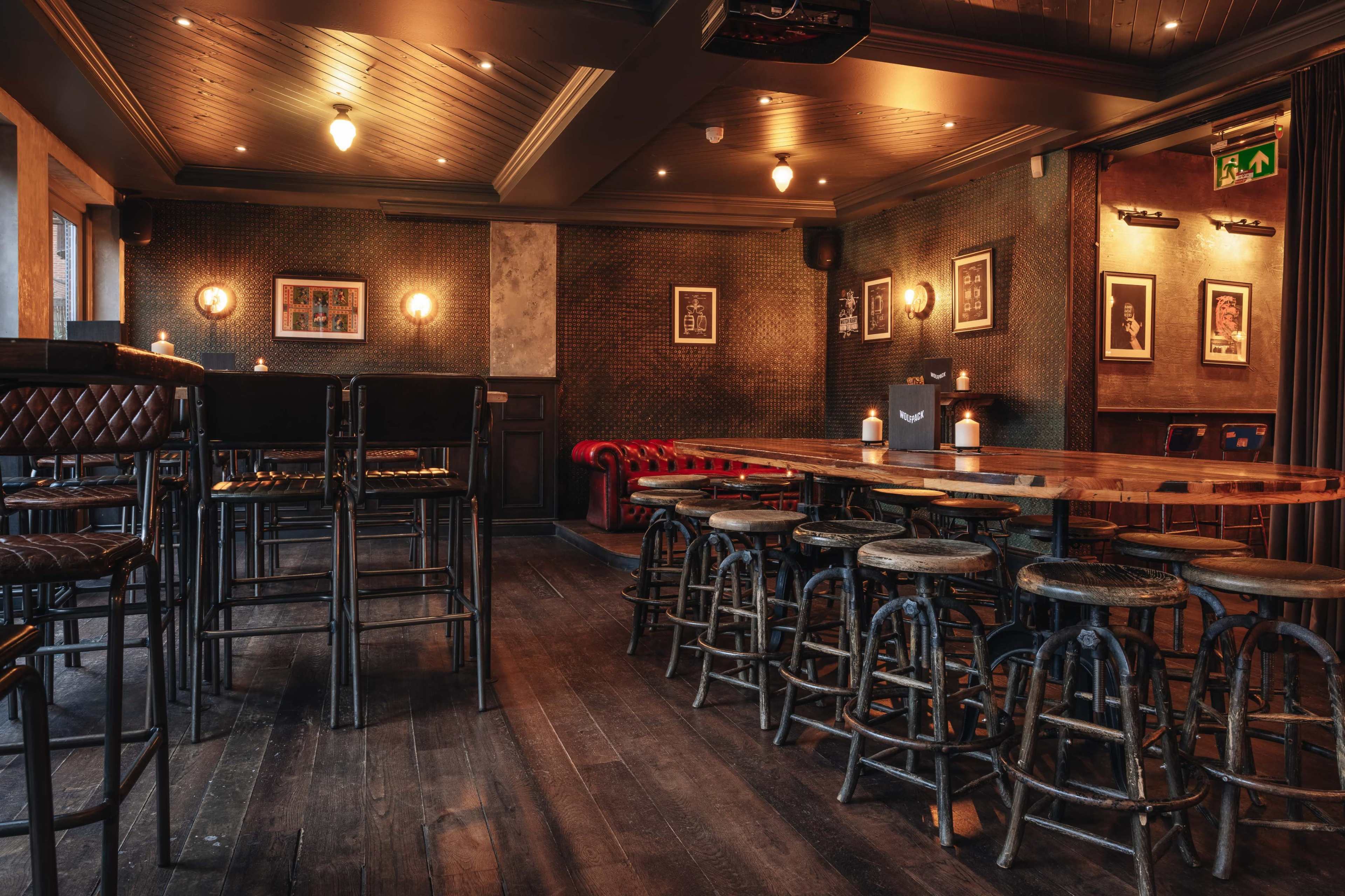 The image shows an empty bar interior with wooden floors, high stools, and a mix of tables and a red leather couch against the wall, decorated with framed pictures.