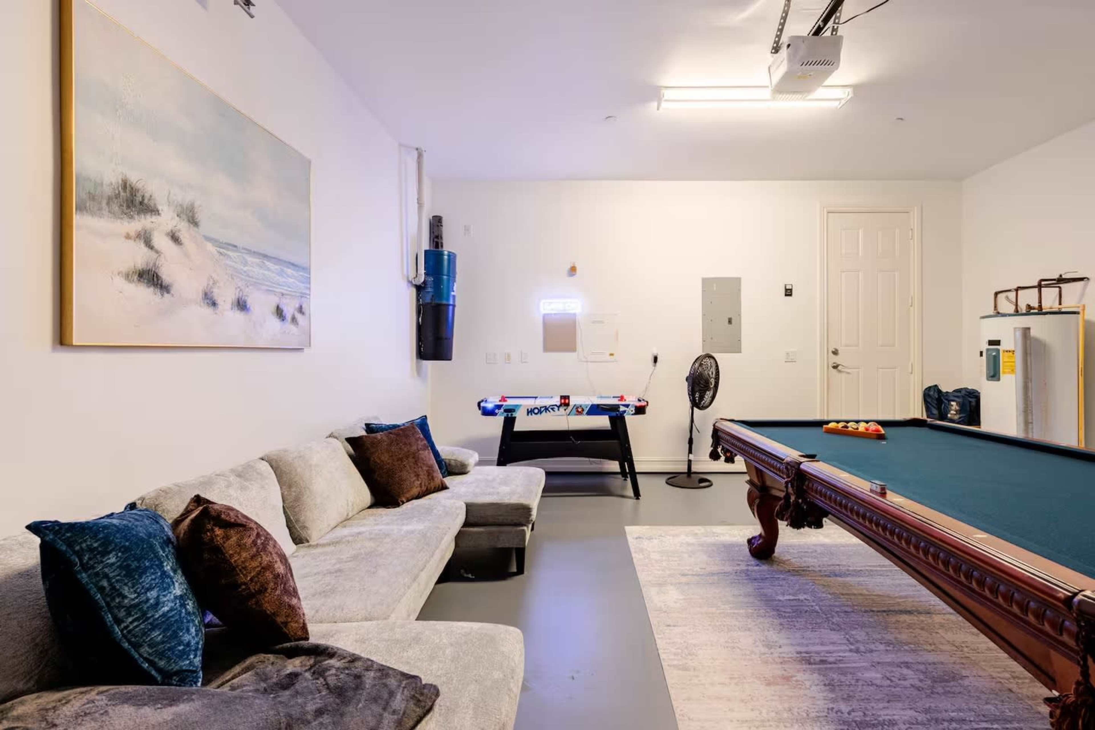 The image shows a garage style room with a pool table, a couch, and an air hockey table, illuminated by artificial lighting.