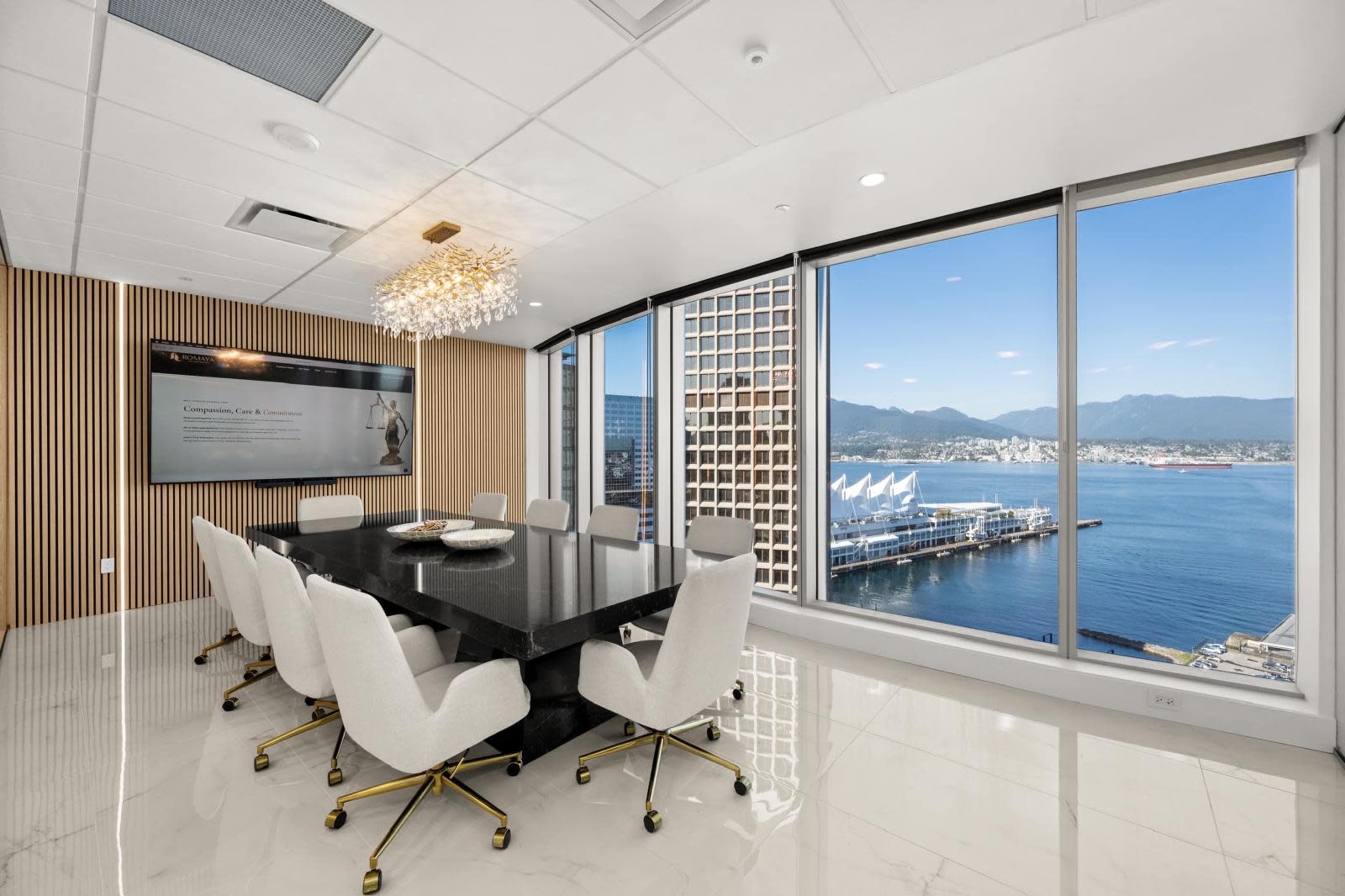 A modern conference room features a large black table surrounded by white chairs, with floor-to-ceiling windows offering a view of a marina and mountains.