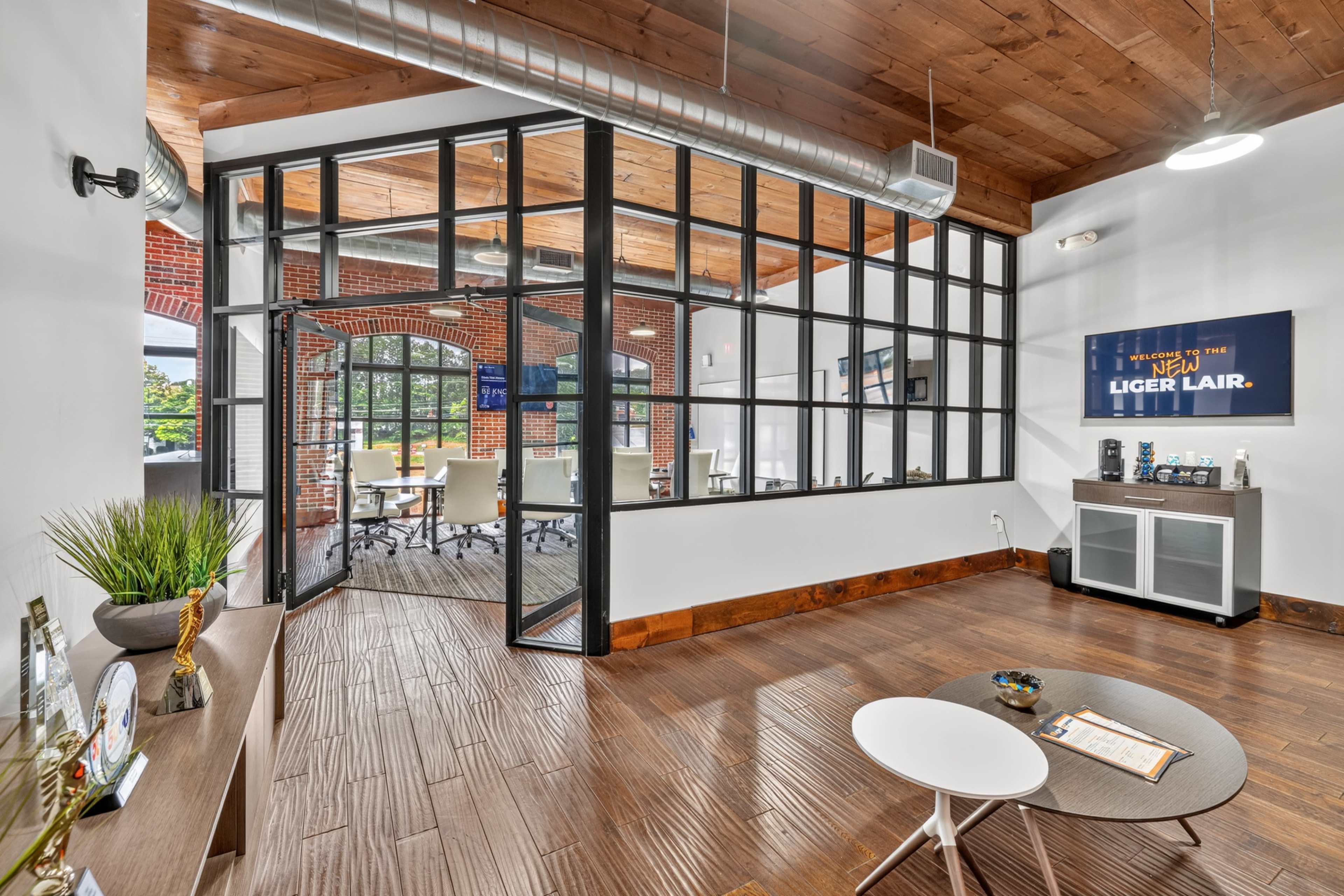 The image shows a modern office space featuring exposed wooden beams, a glass-walled meeting room, and contemporary furniture.