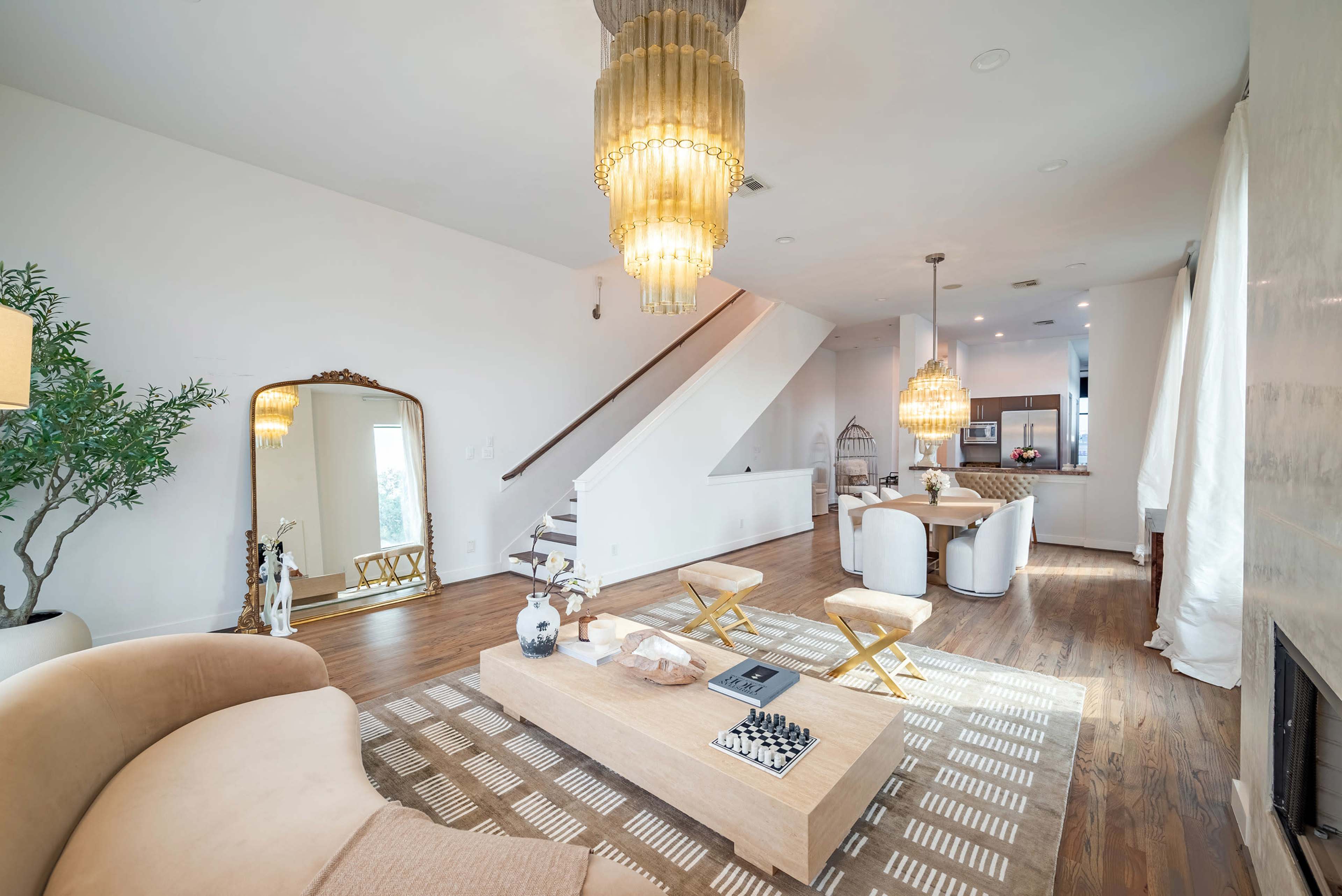 The image shows a modern living room with a staircase, chandelier lights, a large coffee table, and a dining area in the background.