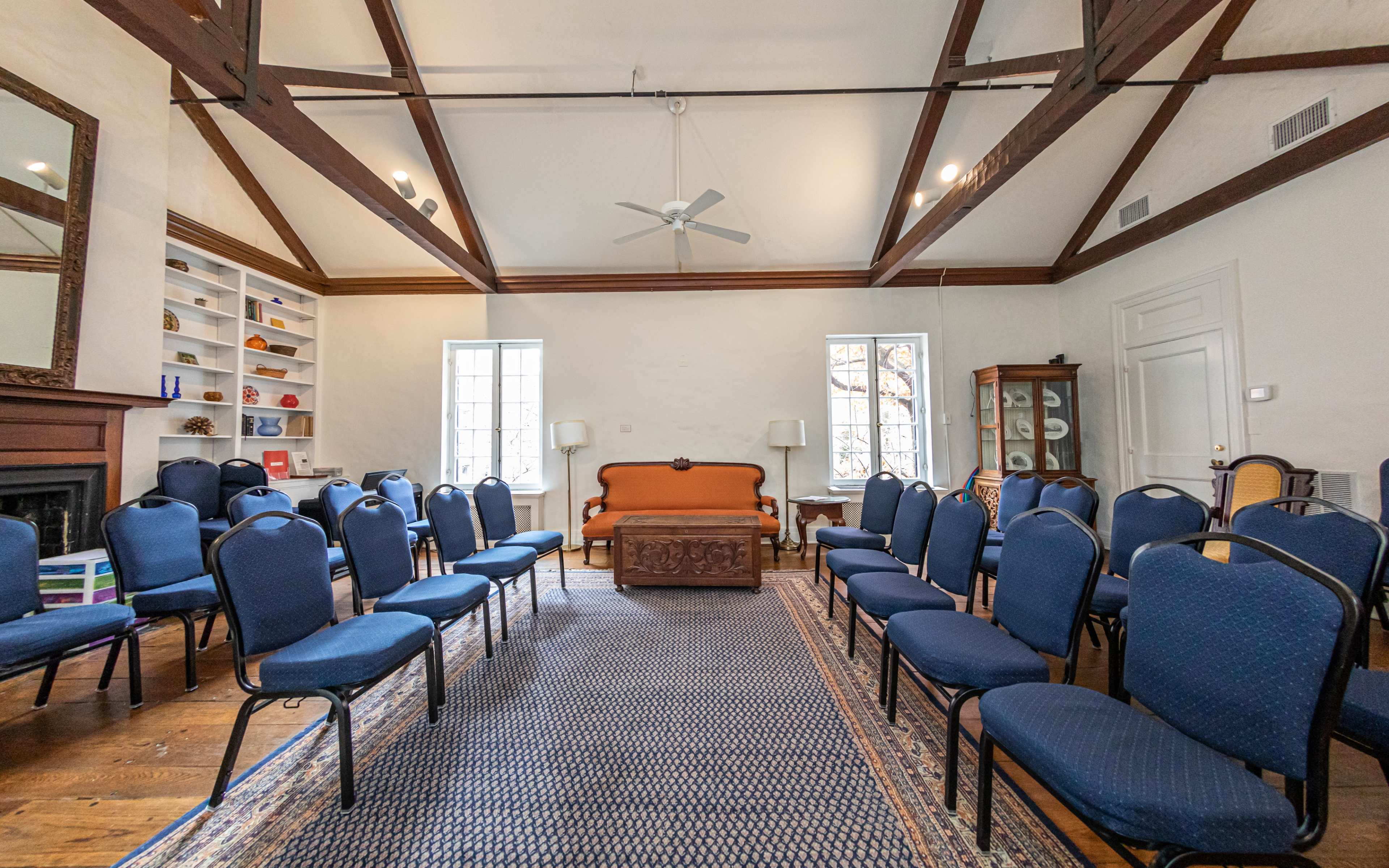 The room features rows of blue chairs facing an orange sofa, with large windows and wooden beam ceilings.