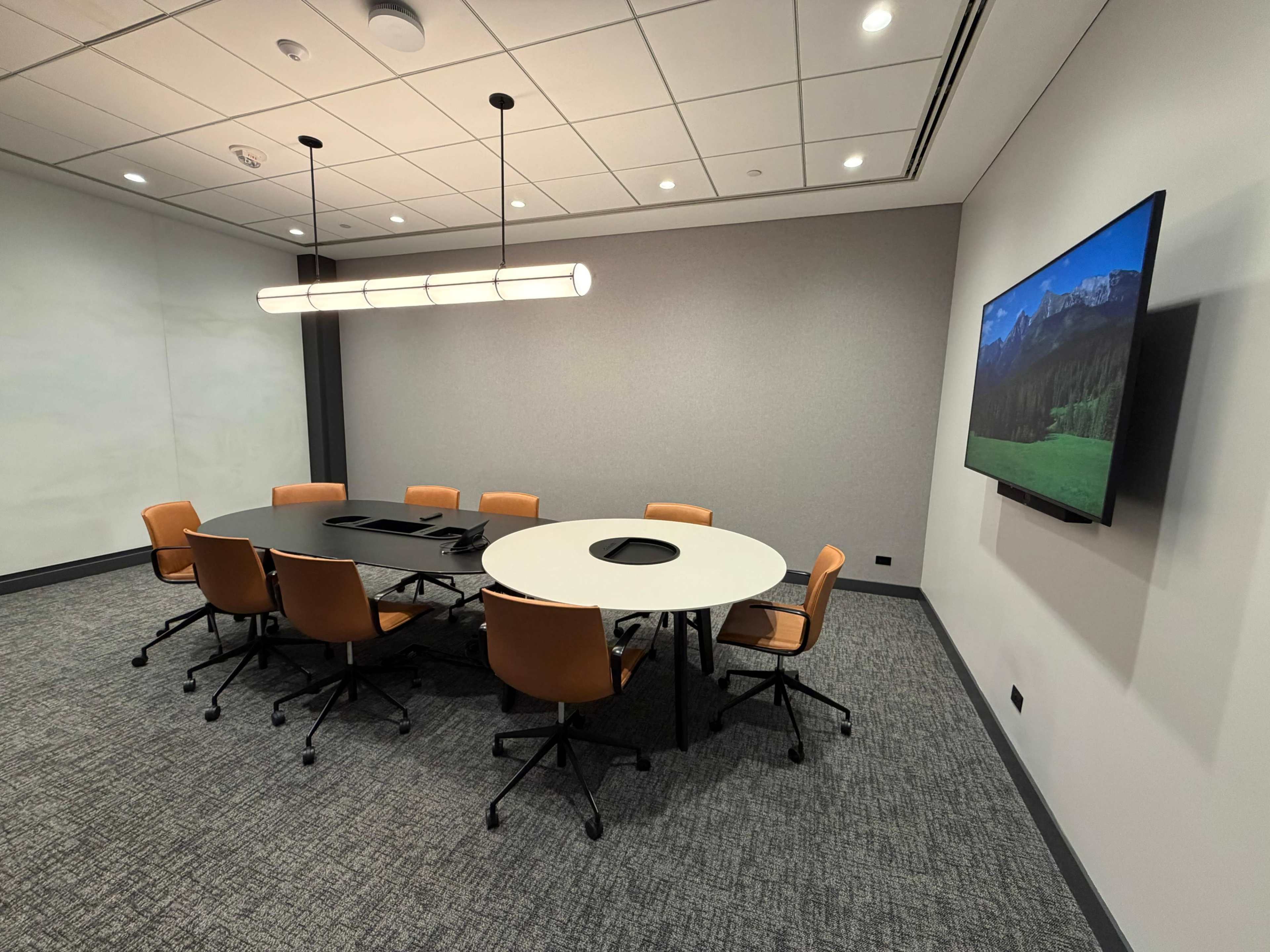 A conference room features a round table surrounded by six brown chairs and a large wall-mounted screen.