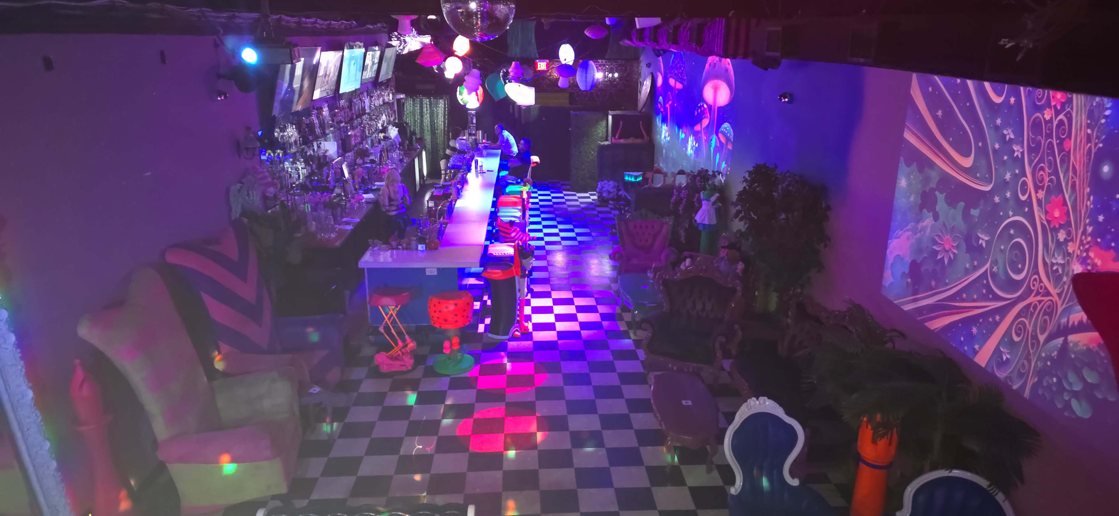 A colorful bar interior with a checkered floor, illuminated by various lights and decorations, featuring a long bar counter and seating areas.