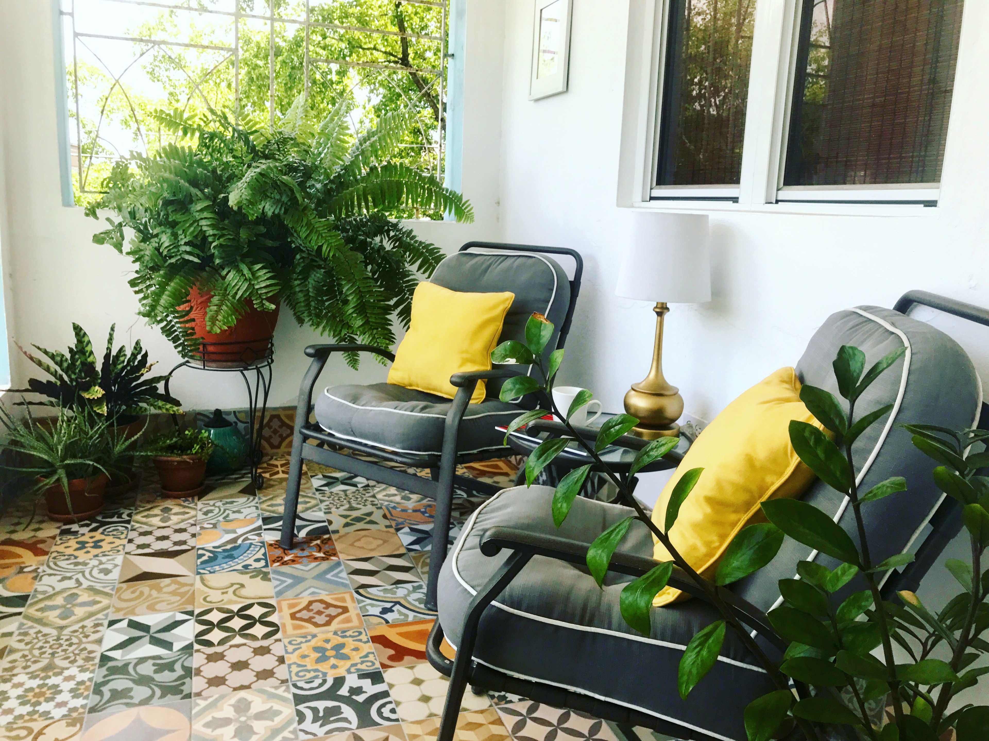 Two gray chairs with yellow cushions sit on a patterned tiled floor, surrounded by various potted plants and a small lamp.