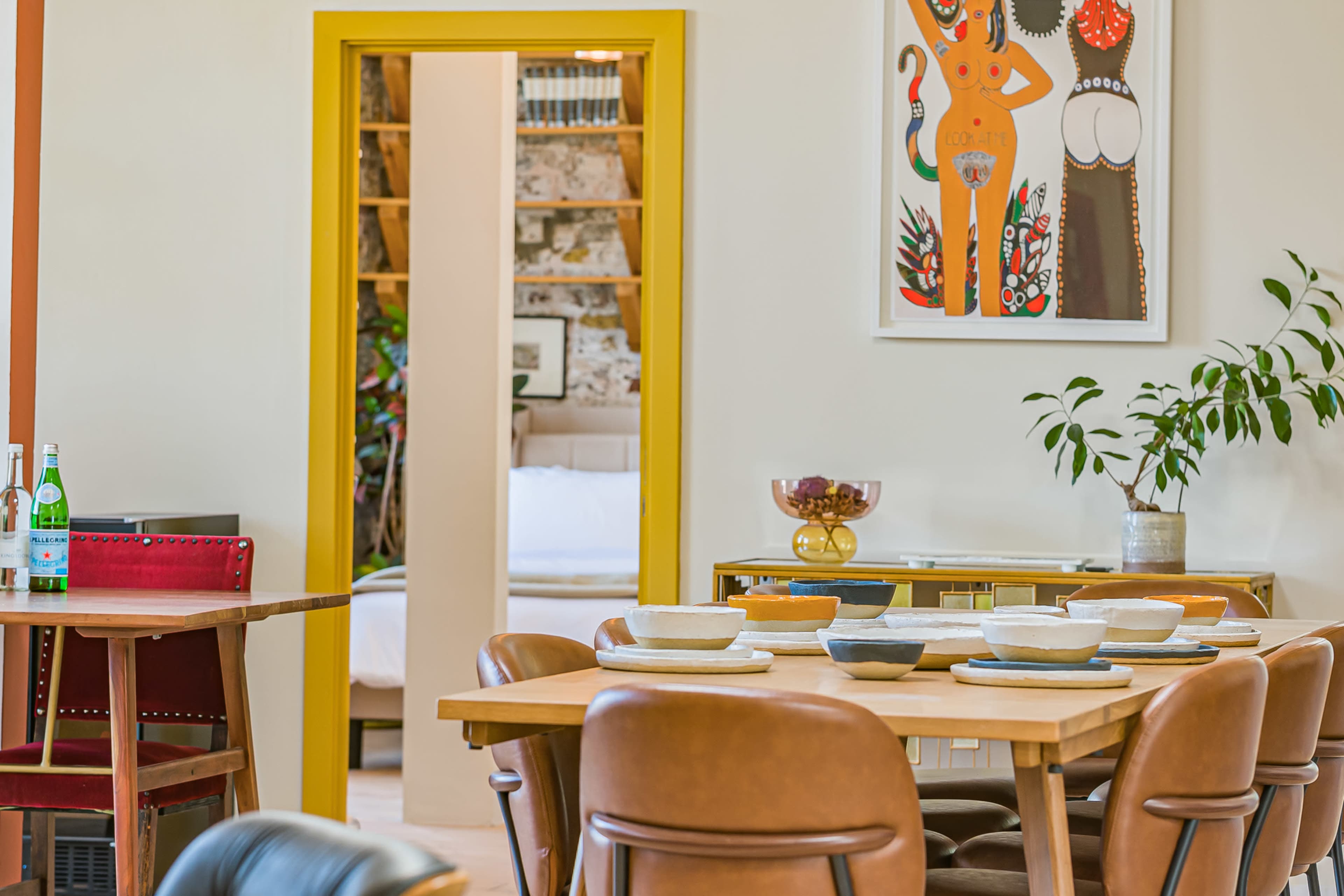 The image shows a dining area with a wooden table surrounded by brown chairs, a colorful artwork on the wall, and a doorway leading to another room in a well-lit space.