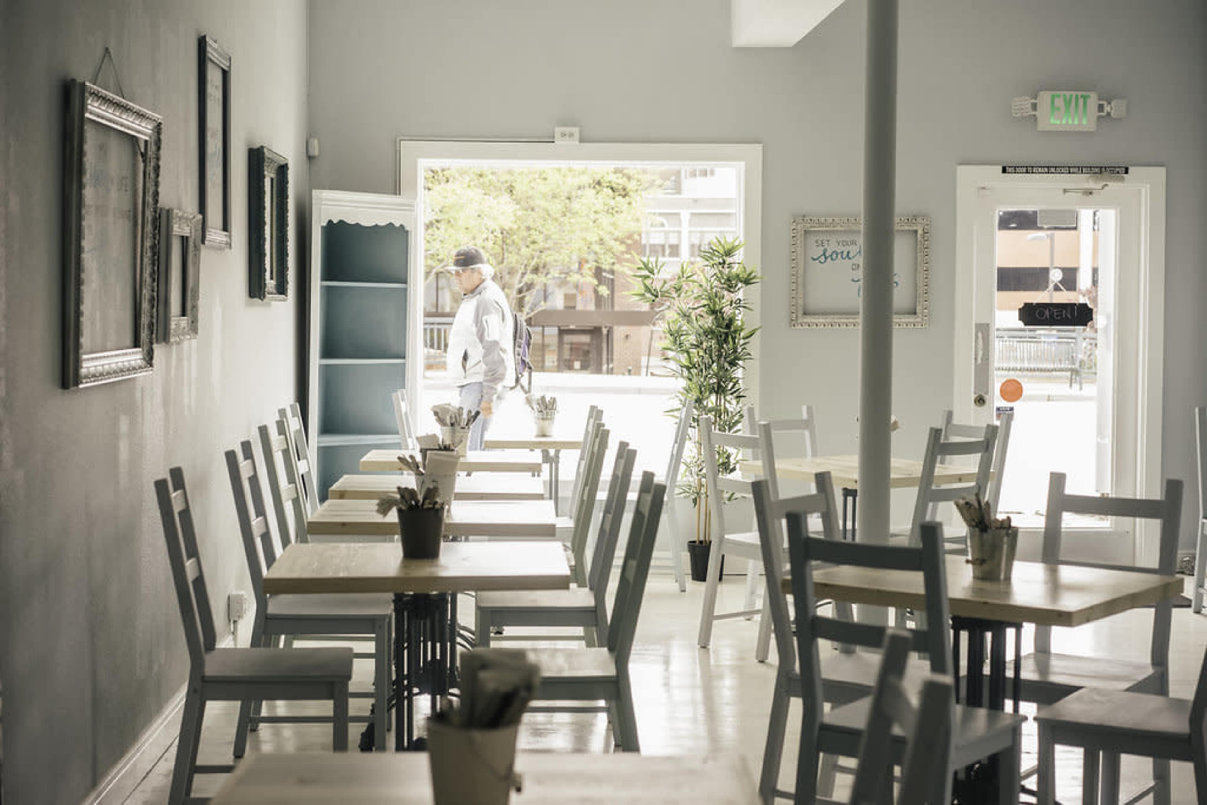 A man enters a light-colored café with empty tables and a view of the outdoor seating area.
