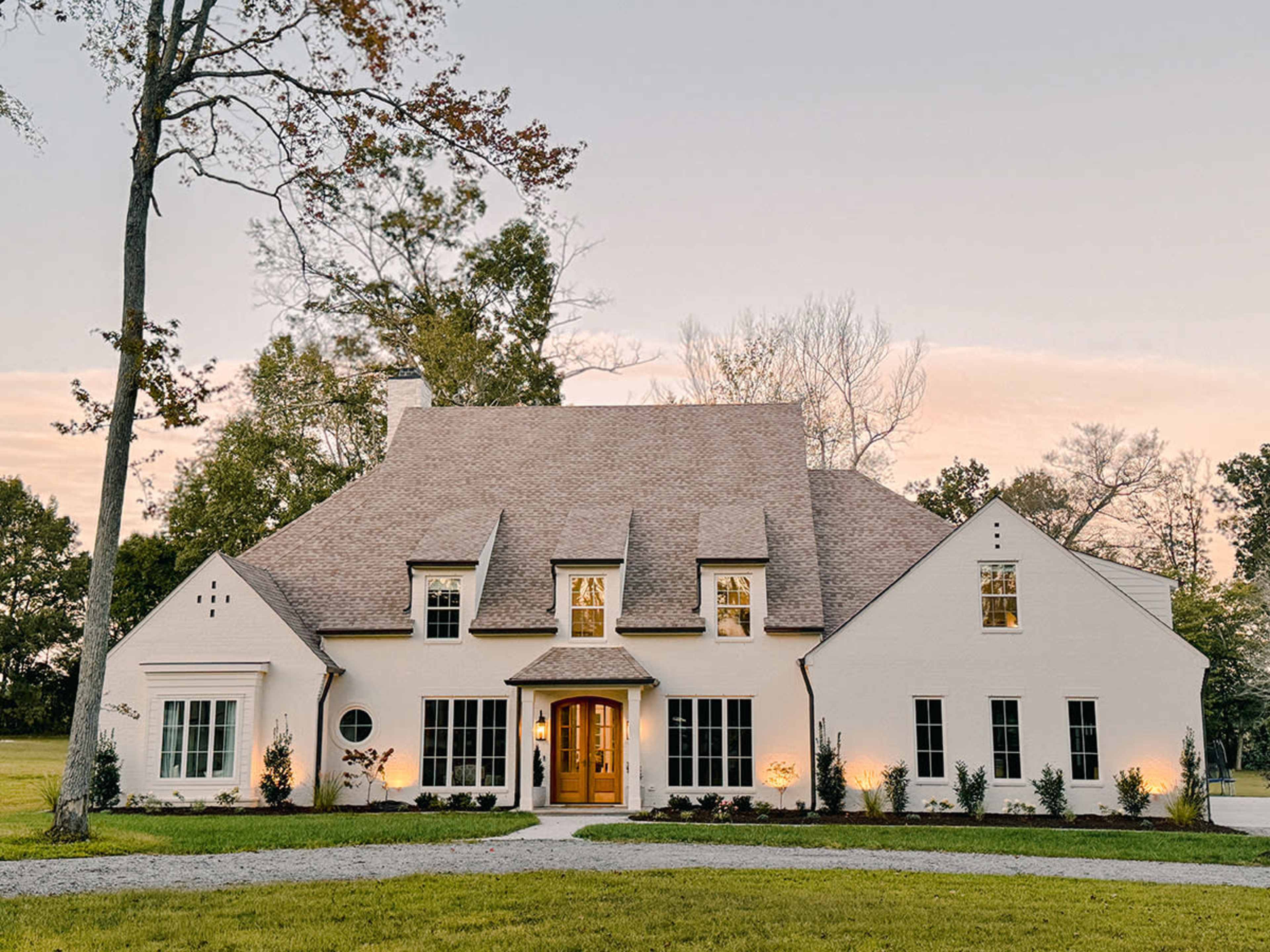 The image shows a white, two-story house with a steeply pitched roof, large windows, and a landscaped front yard.