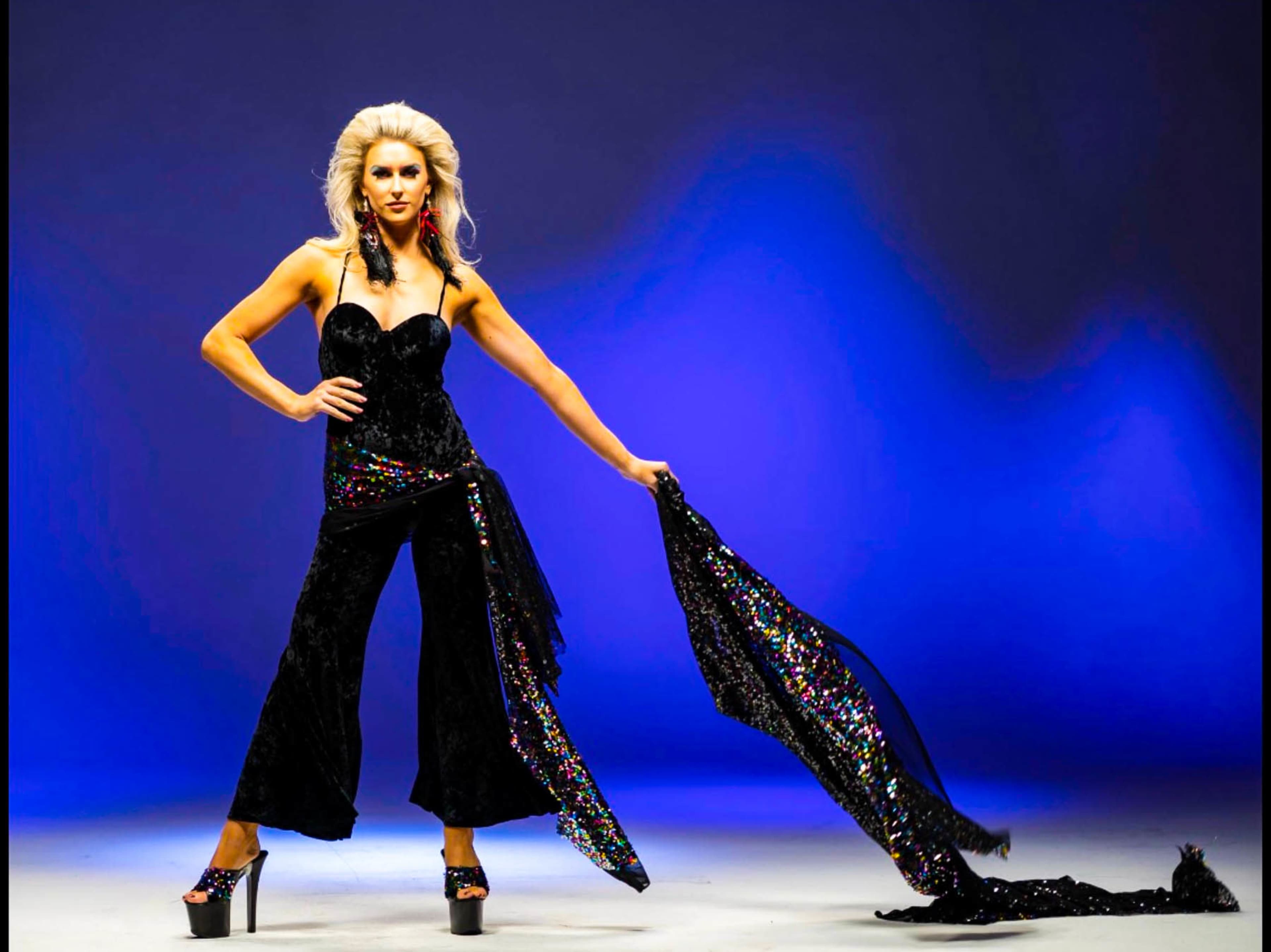 A model poses in a black sequined outfit with flared pants, holding a flowing black cape against a blue backdrop.