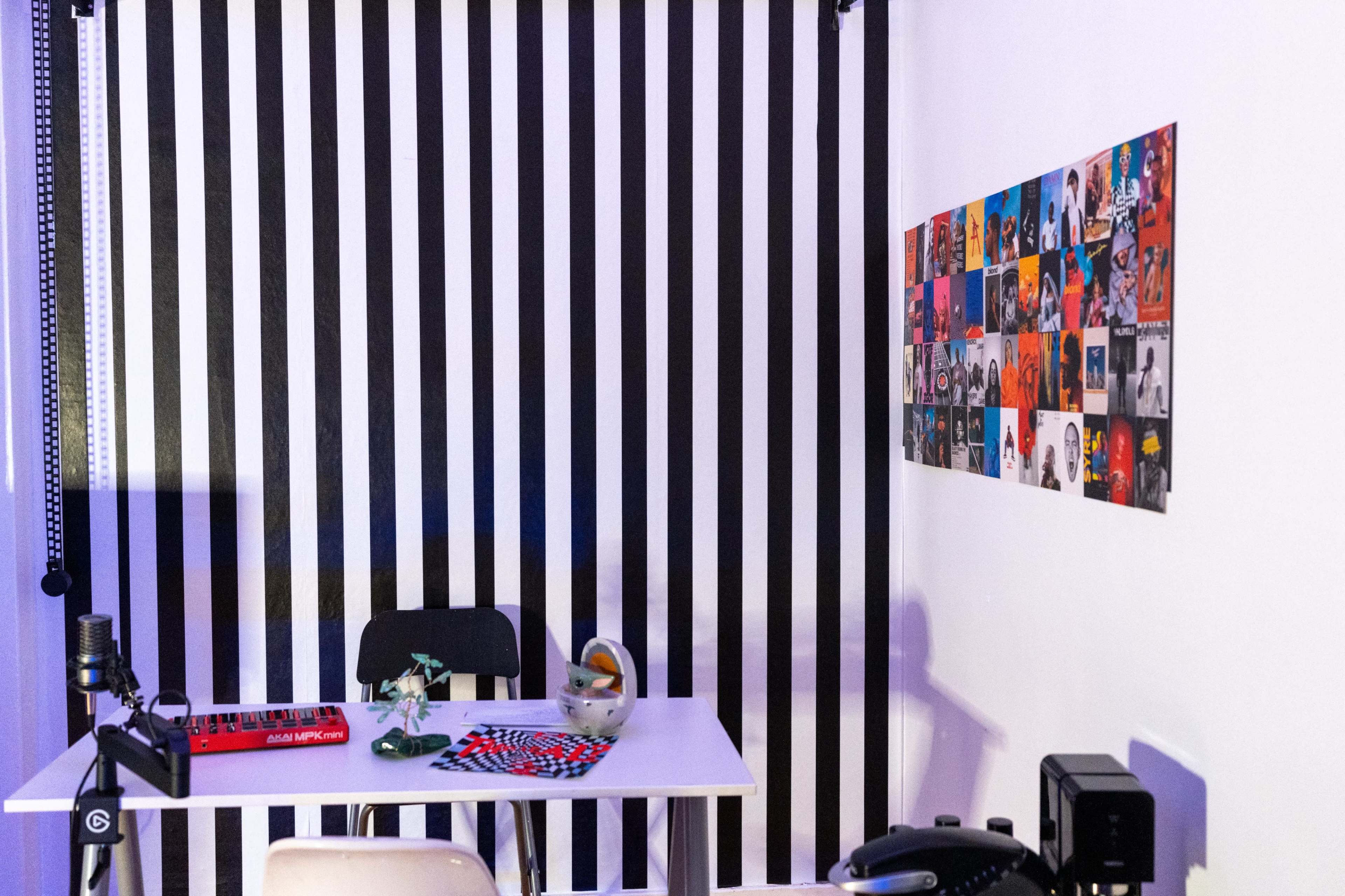 The scene features a room with a black and white striped wall, a white table with a keyboard and a small plant, and a wall adorned with a collage of various colorful images.