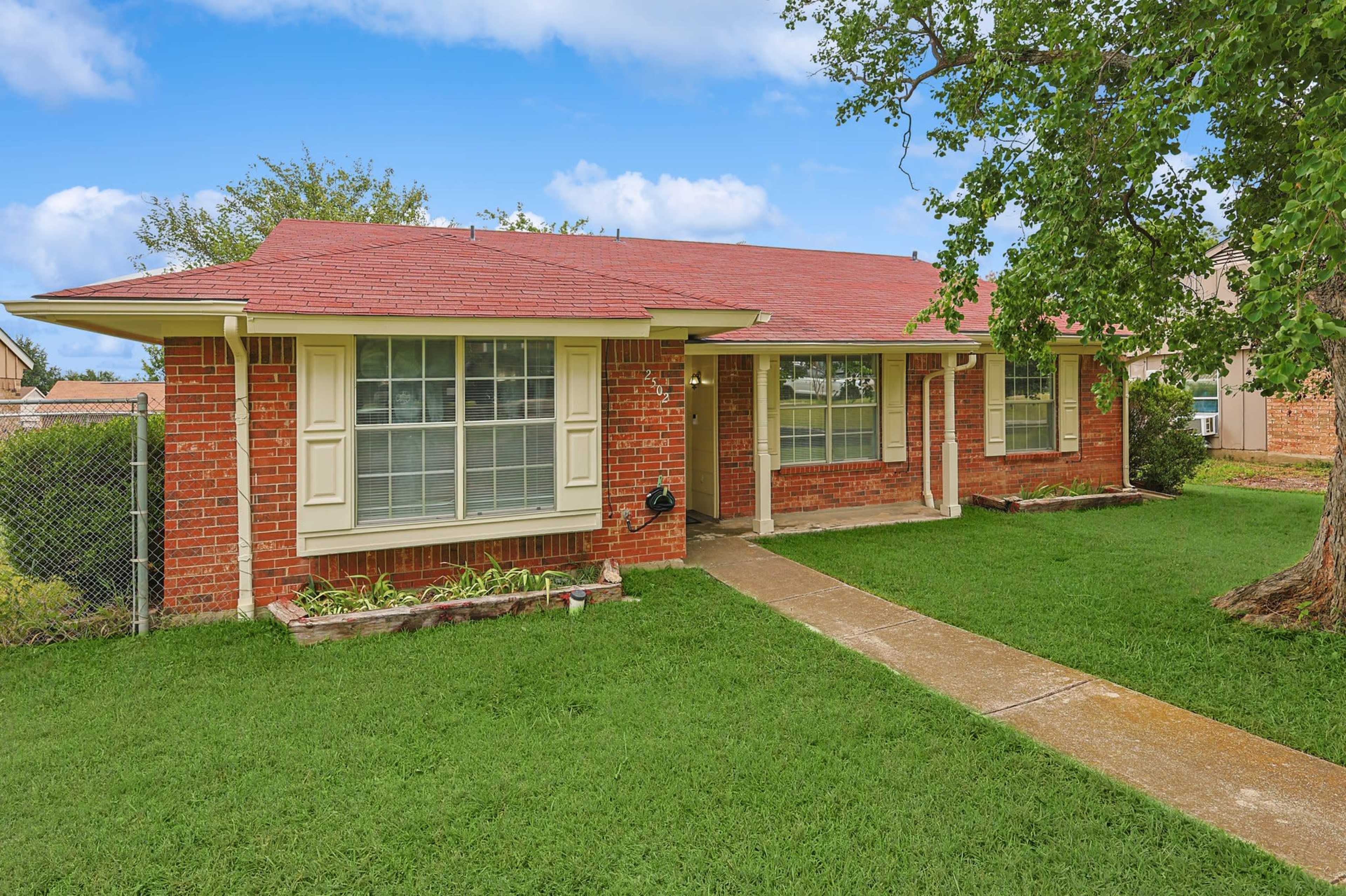 The image shows a single-story brick house with a red roof, large front windows, and a paved walkway leading to the entrance, surrounded by a grassy lawn.