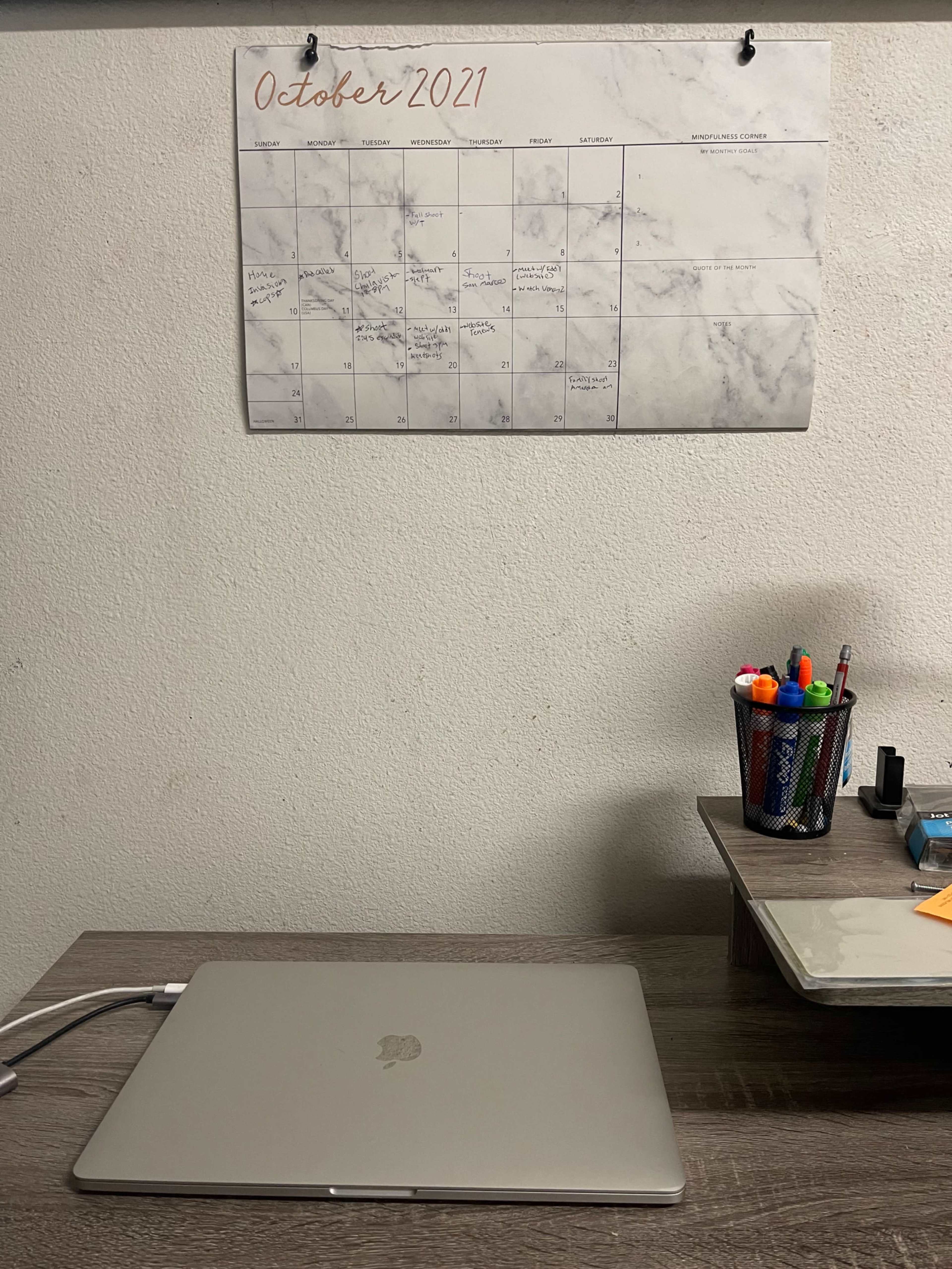 A laptop sits on a wooden desk beneath a wall calendar for October 2021 and a container of writing utensils.