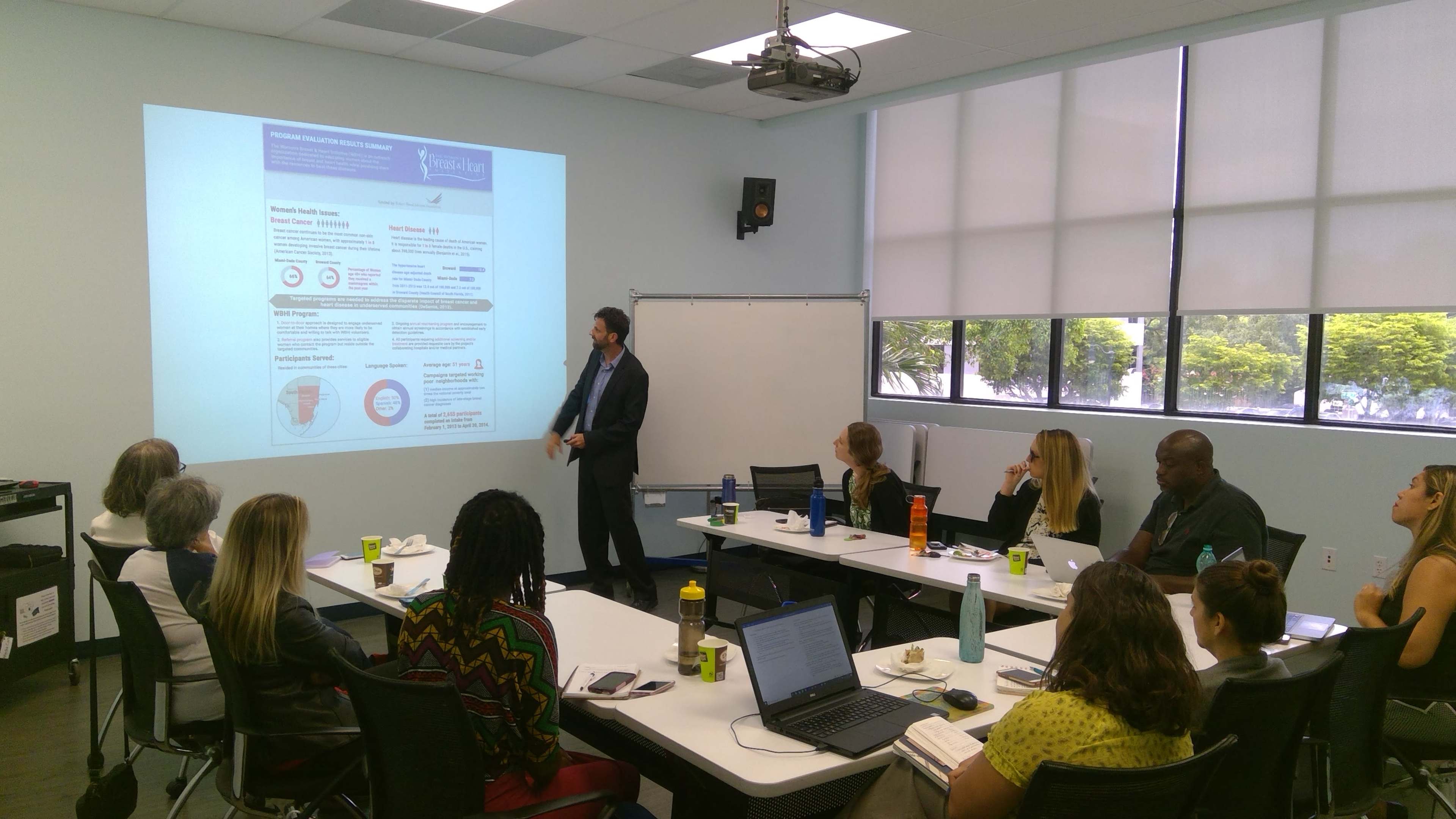 A man is presenting information on a projector to a seated audience in a conference room.